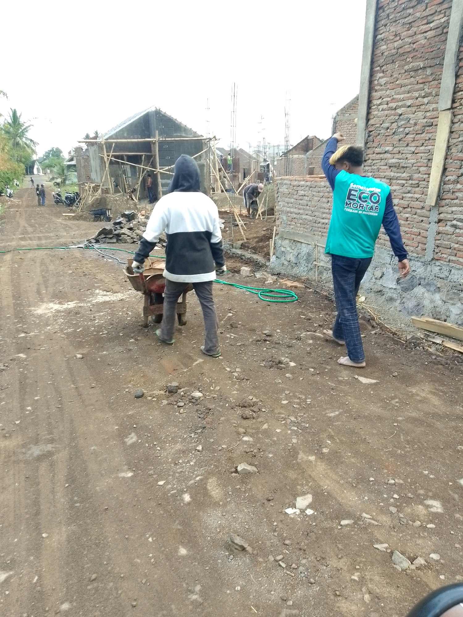 The image depicts a construction site in Jember, Indonesia, where a building is under construction. In the foreground, two men are present. One man, wearing a black and white hooded jacket and gray pants, is pushing a wheelbarrow. The other man, wearing a bright teal shirt with "ECO MORTAR" written on it and blue checkered pants, walks away from the camera, with his right arm raised. Various construction materials and equipment are visible, including scaffolding, bricks, and a green hose. In the background, several other individuals are working on the construction site, and motorcycles are parked nearby. The ground is unpaved and dusty, characteristic of a rural or suburban construction area. The overall scene captures the ongoing work at a construction project in an outdoor setting.