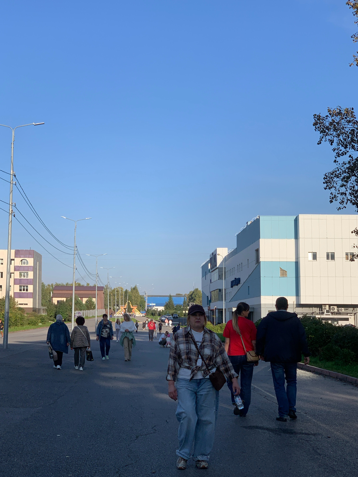 The image depicts an outdoor scene on a paved road in Prokopyevsk, Russia, likely in an urban or commercial area. The sky is clear and blue, indicating fair weather. Several people are walking along the road, some in groups and others individually, moving in various directions. The architecture visible includes modern buildings with light-colored facades, some featuring blue accents, and residential or administrative buildings with purple and beige elements. Streetlights and power lines are present along the road. In the distance, a large, stylized star structure is visible, suggesting a public space or event area. Vegetation, including trees and bushes, lines the sides of the road and surrounds the buildings. The overall atmosphere is one of everyday activity in a city environment.