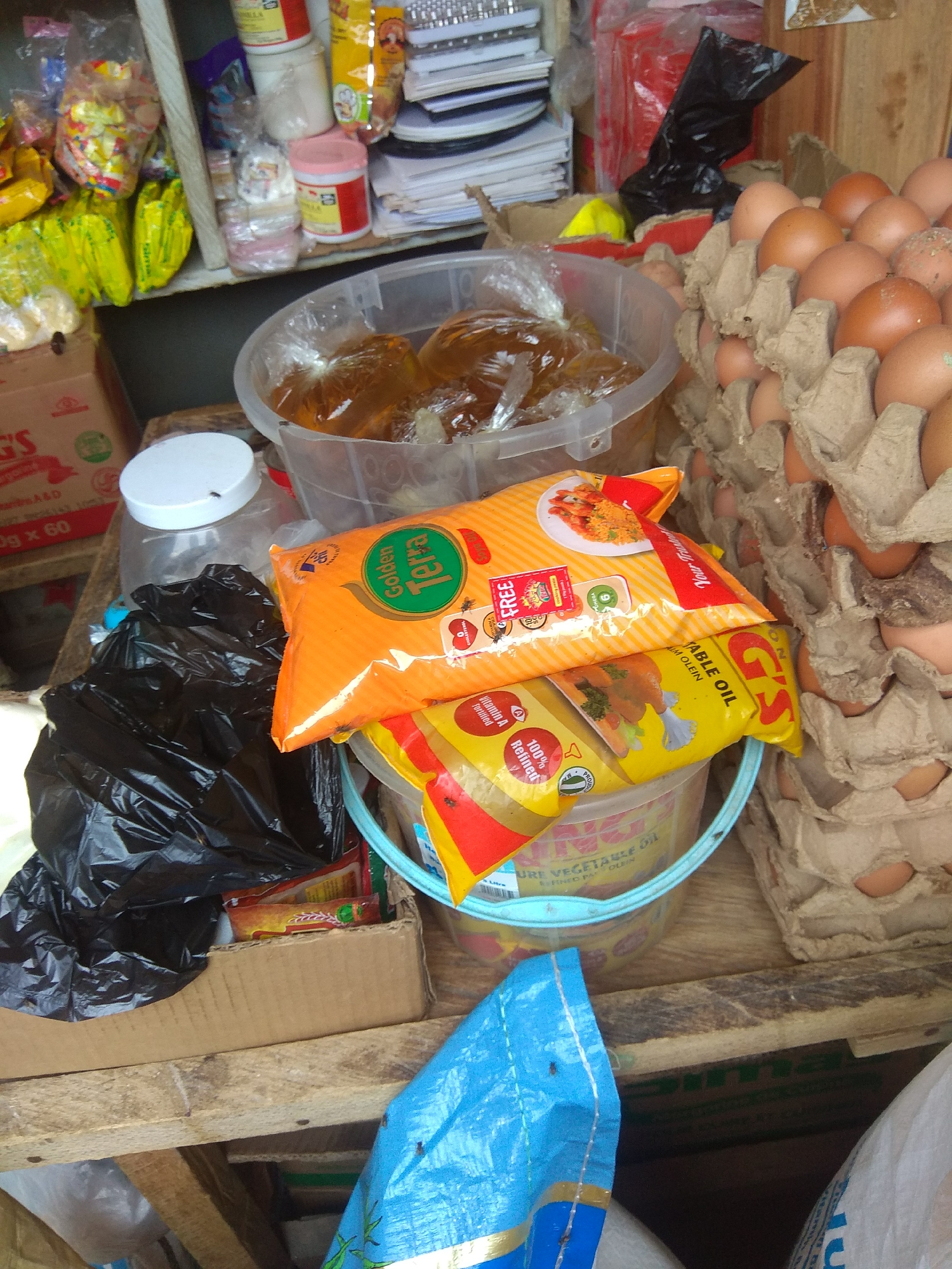 This is an overhead shot of a market stall in Jalingo, Nigeria, cluttered with various food items and supplies. The scene is densely packed with goods, suggesting a busy marketplace. Dominating the foreground are several bags of cooking oil, including prominent yellow bags labeled "G'S PURE VEGETABLE OIL ALM OLEIN" and an orange bag with "Golden Terra" branding. A clear plastic tub filled with a translucent, amber-colored liquid, likely oil or honey, sits near the cooking oil bags.
To the right, a large carton of brown eggs is partially visible, with many eggs nestled in their cardboard packaging. Behind the eggs, a wooden wall with a textured grain serves as the backdrop. In the mid-ground and left, there are more packaged goods, including yellow and clear plastic bags containing what appear to be snacks or other dry food items. A black plastic trash bag is also visible, adding to the overall disarray. Further left and slightly behind, a white plastic container with a lid, possibly for spices or grains, is present.
The lighting suggests it is daytime, with no strong shadows indicating a specific time. The overall impression is one of a working market, with goods arranged for sale. The presence of familiar food packaging brands like "G'S" and "Golden Terra" provides specific detail. The image captures the everyday commerce of a Nigerian market.