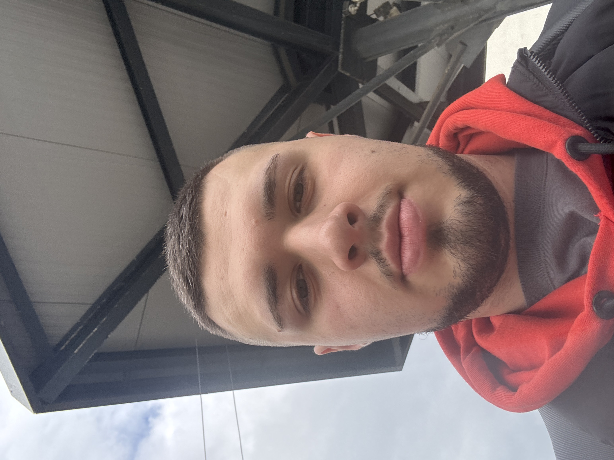 A man is pictured in a close-up selfie, taken from a low angle. He has short dark hair and a beard. He is wearing a red hoodie under a dark-colored jacket. The background shows a metallic structure, possibly a roof or a part of a building's framework, with a cloudy sky visible beyond. The scene could be within an urban environment but there is insufficient detail to place him in Sofia, Bulgaria with certainty.