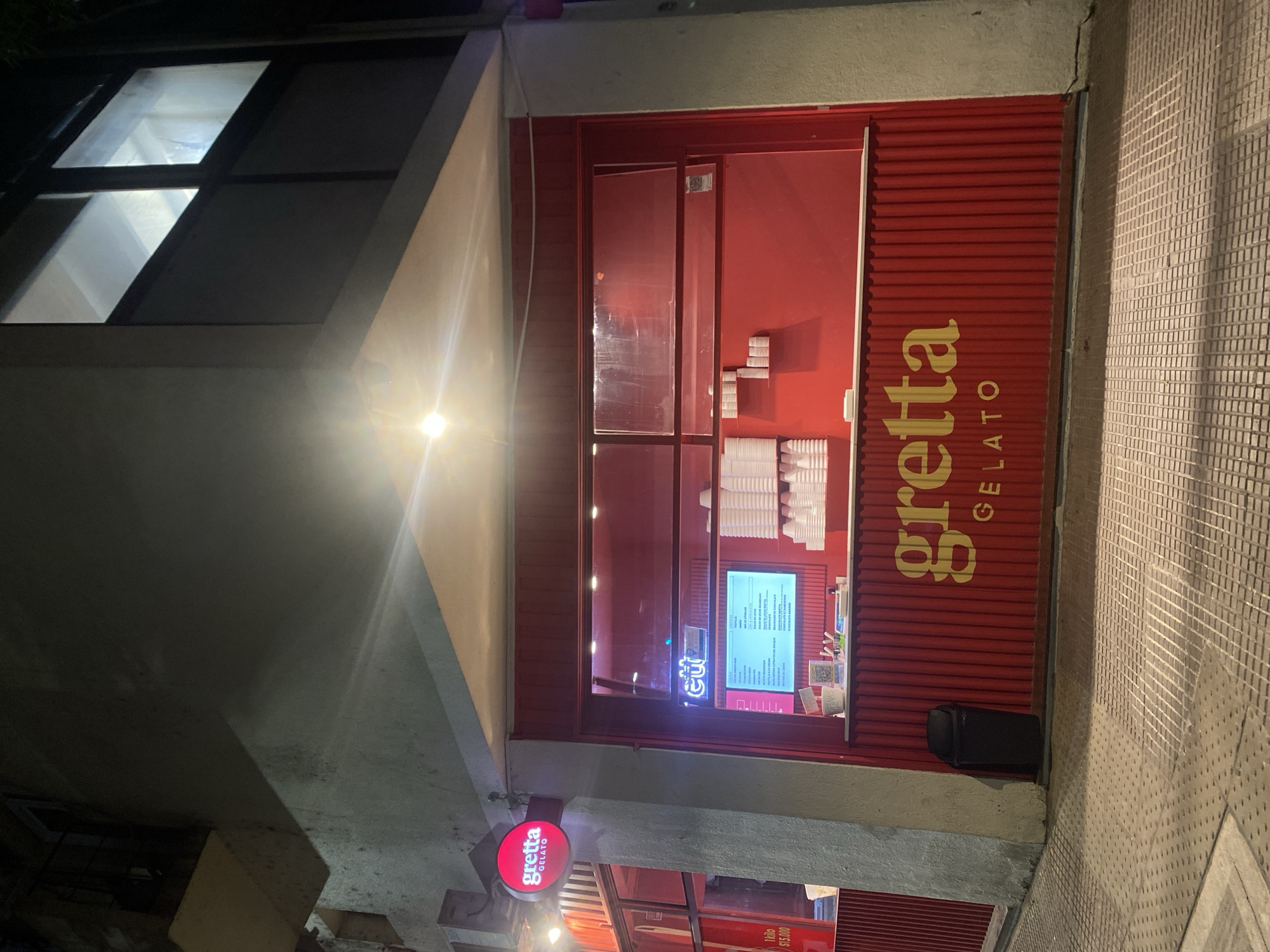 The image displays a nighttime exterior view of a "Gretta Gelato" shop situated on a street corner in Buenos Aires, Argentina. The building, constructed of light-colored concrete or stucco, features a vibrant red facade for the ground-floor gelato establishment. A circular red sign with "gretta GELATO" in white lettering is mounted on the upper left corner of the shop's entrance. The main service area consists of a wide, open window or counter protected by a transparent glass pane, through which the shop's interior is visible. Inside, a brightly illuminated digital menu board displays a list of gelato flavors, and numerous stacks of white plastic cups are arranged on shelves and the counter. The lower section of the shop's red facade is adorned with vertical corrugated paneling, featuring "gretta GELATO" in large yellow text. To the far left, a smaller red sign indicates "1 kilo $15.000". A black, lidded waste bin rests adjacent to the red facade near the corner. The foreground reveals a patterned tiled sidewalk, characteristic of Buenos Aires urban environments, illuminated by the shop's interior lights and an overhead fixture. The upper levels of the building exhibit windows, some with internal illumination. No individuals, actions, or interactions are present in the depicted scene.
