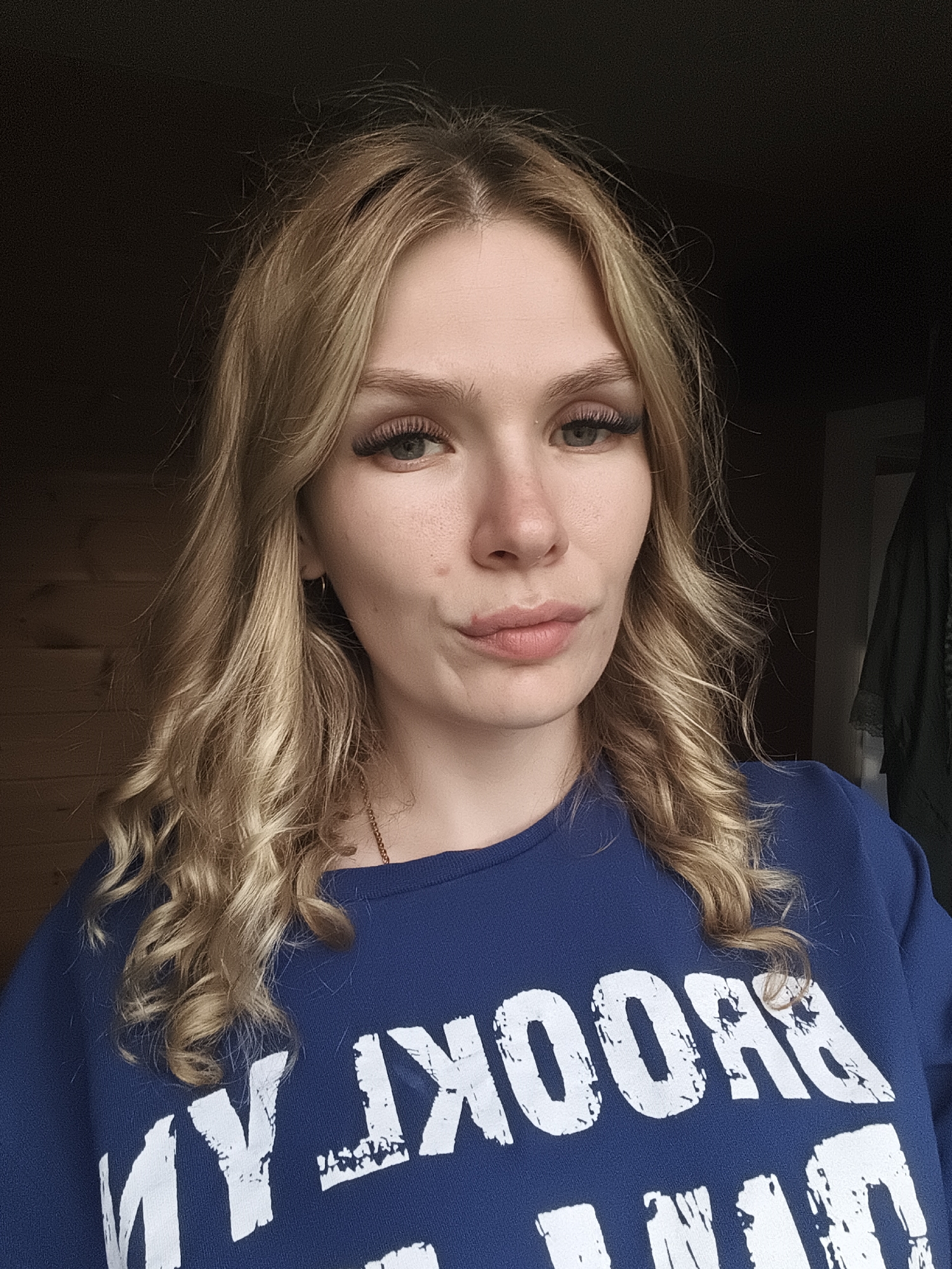 A young woman with light blonde, shoulder-length, wavy hair, and light eyes looks directly at the camera with a slight pout. She is wearing a dark blue sweatshirt with white text that reads "BROOKLYN." The background appears to be an interior setting, with a dark wooden wall on the left and a dark doorway with a garment hanging behind her on the right. The location, Русский Вожой, Russia, cannot be determined from the image.