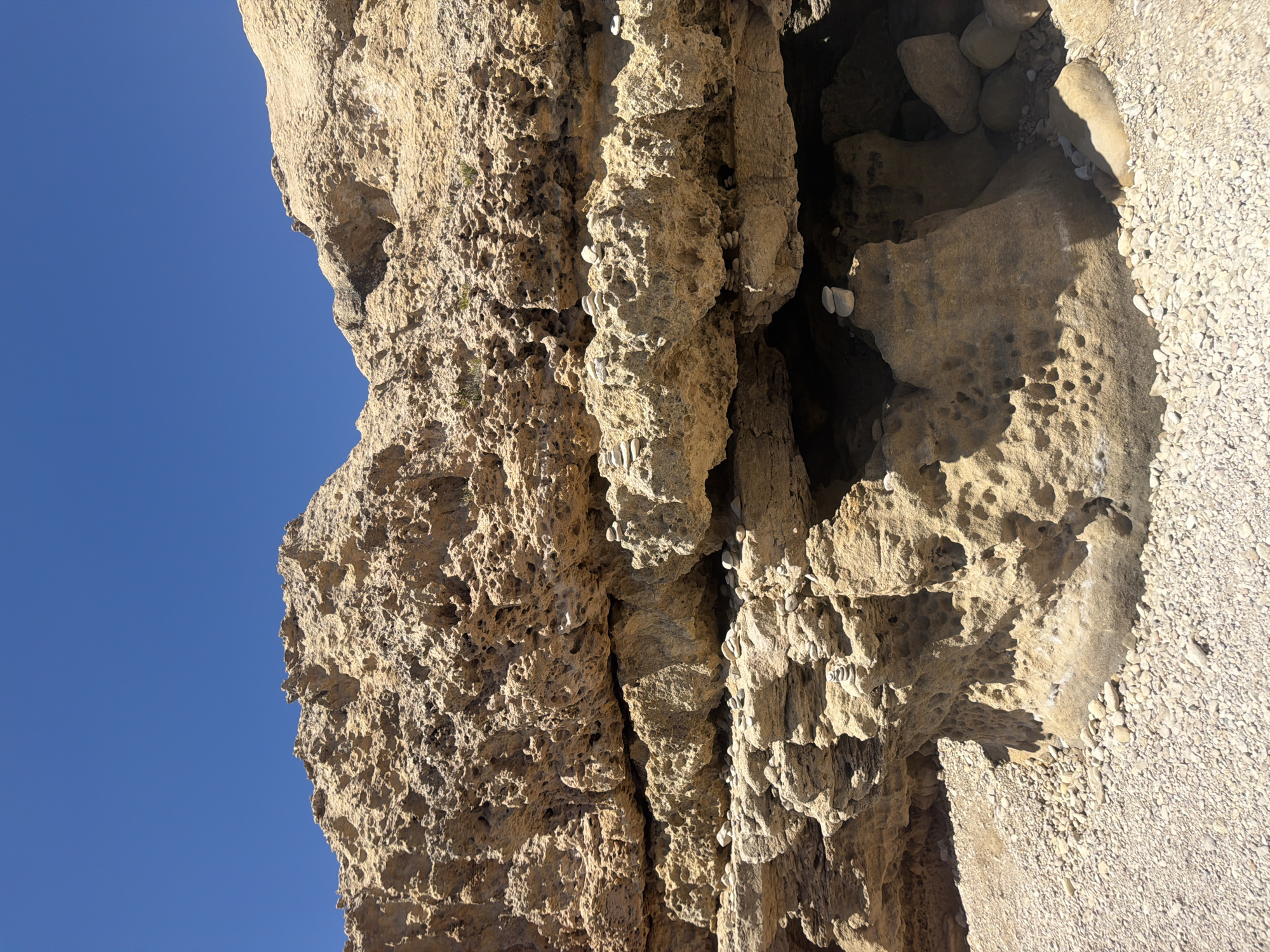 The image shows a natural rock formation under a clear blue sky. The rocks are tan and textured, with visible layers and small holes, suggesting erosion. In several places, small stacks of pebbles have been carefully arranged on ledges and within the rock crevices. The ground in the foreground is covered with small, light-colored pebbles. The overall scene appears to be a coastal or seaside environment. The specific location in Saint Lawrence, Malta, is not identifiable from the image alone.