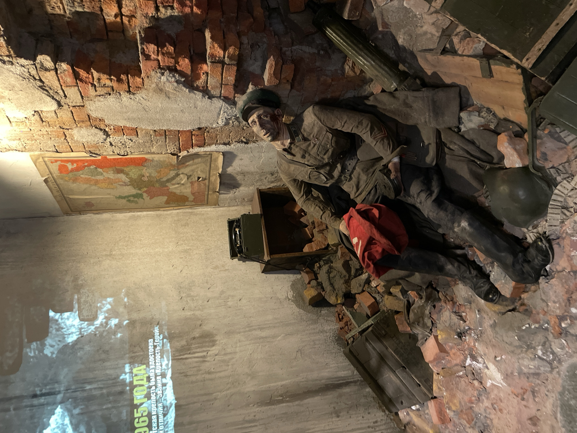 Here is a description of the image.
The image shows a diorama of a fallen Soviet soldier within a brick-walled ruin, likely meant to represent a scene from World War II. The soldier is dressed in a period-accurate uniform, complete with boots, and has a red bag nearby. He has a dark colored head cover on. Nearby, there is a map affixed to the wall and a field telephone unit. The scene is set indoors, and the angle is tilted to the right. A green, tube-like object and a helmet lie near the soldier. The walls appear to be constructed of damaged red brick and crumbling plaster. Text is visible on the lower left of the wall, but the specific content isn't clear. The diorama depicts a scene of conflict and destruction. The diorama is located in the Museum of the Great Patriotic War in Moscow, Russia.