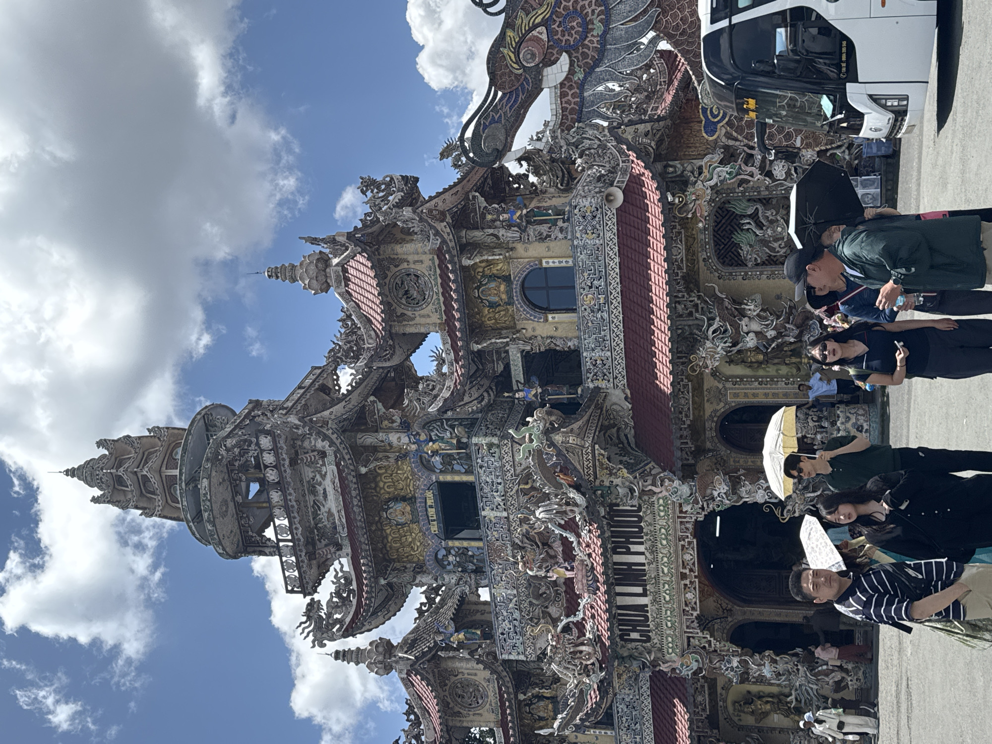 In Xuân Thành, Vietnam, a large, ornate building, likely a temple, dominates the scene under a bright, cloudy sky. The architecture features intricate, colorful decorations, including dragon motifs and numerous statues, with a prominent sign reading "CHUA LINH PHUOC." In the foreground, a group of people stands looking towards the building. A man in a striped shirt is on the left, a woman in a black top is next to him, and a man in a green jacket holds a black umbrella. To the right, a white tour bus is parked. The ground is paved, and the overall impression is of a vibrant tourist destination.