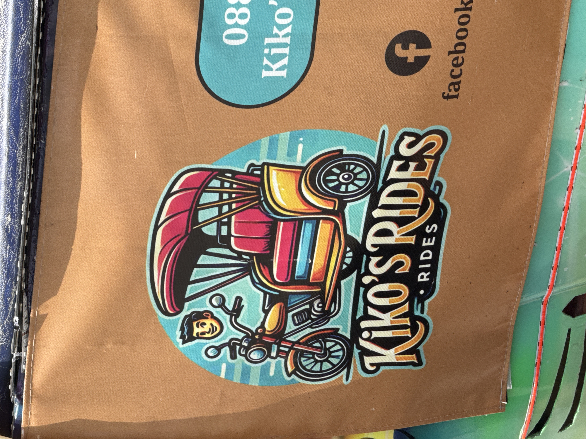 This close-up image displays a bright and colorful illustration on a light brown, fabric-like surface, possibly a bag, sign, or cover, set in Varna, Bulgaria. The central subject is a stylized, motorized pedicab or tuk-tuk, rendered in vibrant reds, yellows, blues, and oranges, with prominent black outlines. A small, cheerful cartoon character's head, presumably the driver, is depicted to the left of the vehicle.
The activity suggested is that of providing rides or transportation services. The illustration is placed within a light blue circular background with subtle vertical stripe patterns.
Notable details include the clear, bright lighting suggesting daytime. The aesthetic is playful and inviting, with the cartoon driver having a simple, happy expression. To the left, a dark blue, textured surface with visible stitching, possibly a binder or another part of an item, borders the brown material. To the right, a partially visible green or teal surface with a red line indicates another object or part of the environment.
Visible text on the brown surface includes "Kiko's RIDES" in a large, stylized font, with "RIDES" smaller and beneath "Kiko's". Above the main illustration, a light blue oval contains the partially visible text "088" and "Kiko'", likely part of a phone number and the name of the business. Further up and to the right, a Facebook logo is accompanied by the word "facebook".