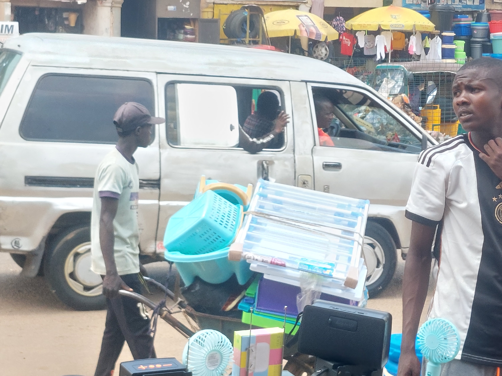 A light-colored van is parked on a dirt road in front of stalls selling various items, including clothing, buckets, and what appear to be household goods. A young man wearing a cap and a light-colored t-shirt is walking in front of the van, pushing a cart laden with blue plastic baskets, a folding rack, and other items. A man in a black and white t-shirt stands to the right of the van, looking towards the right. Two people are visible inside the van, one in the driver's seat and another leaning out of the passenger window. The scene appears to be a street market in an urban or semi-urban area.