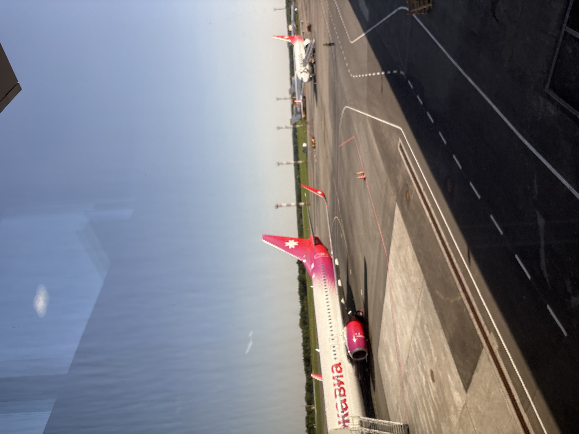 A sideways, medium-angle view of an airfield in Новое Мартьяново, Russia. Two parked aircrafts are visible. The aircraft closest to the viewer is predominantly white with pink accents on its tail fin and engine cover. The words "NORDWIND" are written along the length of the aircraft, in reverse due to the window it is viewed from. In the distance, another aircraft is parked, also painted white, with a red tail fin. The airfield surface is dark grey and marked with white and red lines. Part of a building, possibly the airport terminal, is seen on the left side of the image, where the image has the appearance of viewing through glass. The sky is light blue with a greyish hue towards the horizon. The photograph appears to be taken from inside a building looking out onto the airfield.
