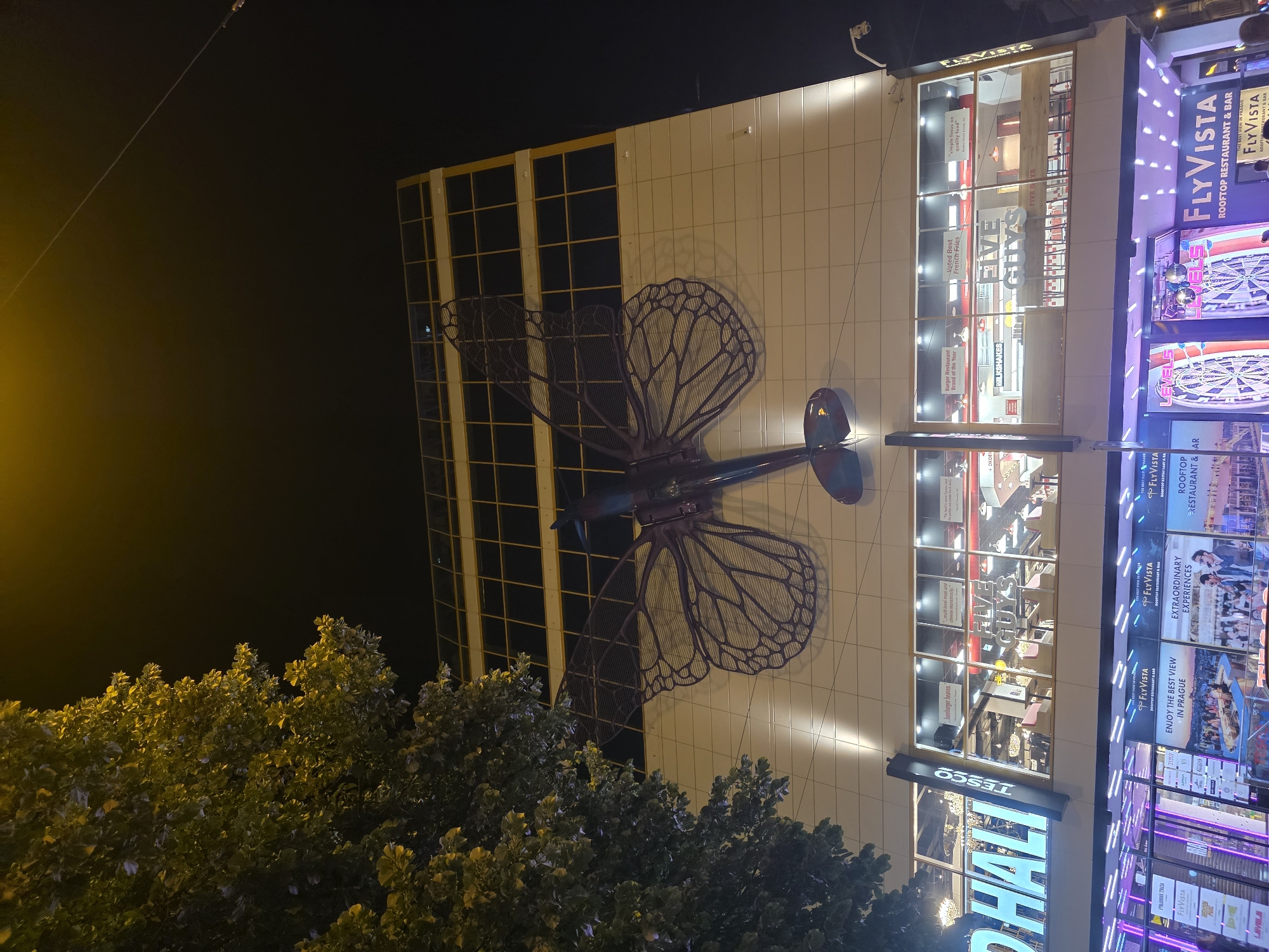 This is an outdoor nighttime shot of the FlyVista Rooftop Restaurant and Bar and Tesco storefronts in Prague, Czech Republic. The image includes a golden building structure with black windows, a cream-colored building structure with a large, metallic, steampunk dragonfly sculpture attached to it, the Five Guys restaurant, signage with the word "Flyvista" written on it, and trees.