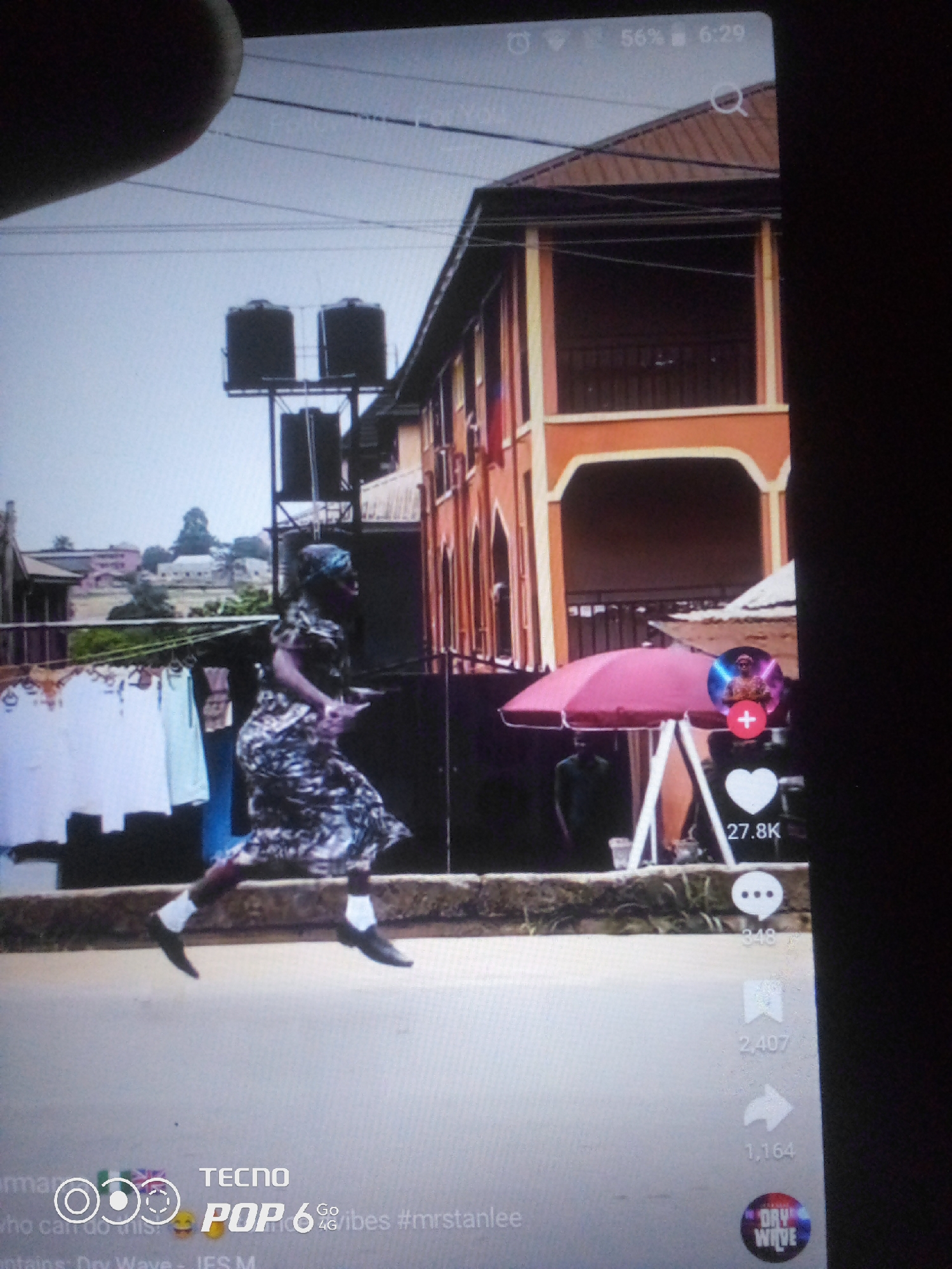 The image depicts an outdoor scene in the city of Howayi, Nigeria, with a focus on a woman in mid-stride. The woman, dressed in a patterned dress and headscarf, is captured in motion, her legs extended as if running or dancing. She is positioned in the foreground, with a street and a low wall behind her.
In the background, a multi-story building with an orange facade and balconies dominates the scene. To the left of the building, clothing is seen hanging on a clothesline, suggesting a residential area. Two large water tanks are mounted on a structure near the building. A pink umbrella stands open near the building's entrance, with a person partially visible beneath it.
The lower portion of the image shows overlaid graphics from a mobile application, including icons for likes, comments, shares, and a profile picture with a "plus" icon. Text indicating "TECNO POP 6", "Go", "4G/vibes #mrstanlee", "Dry Wave", and numerical counts for engagement metrics are also present. The overall lighting suggests it is daytime.