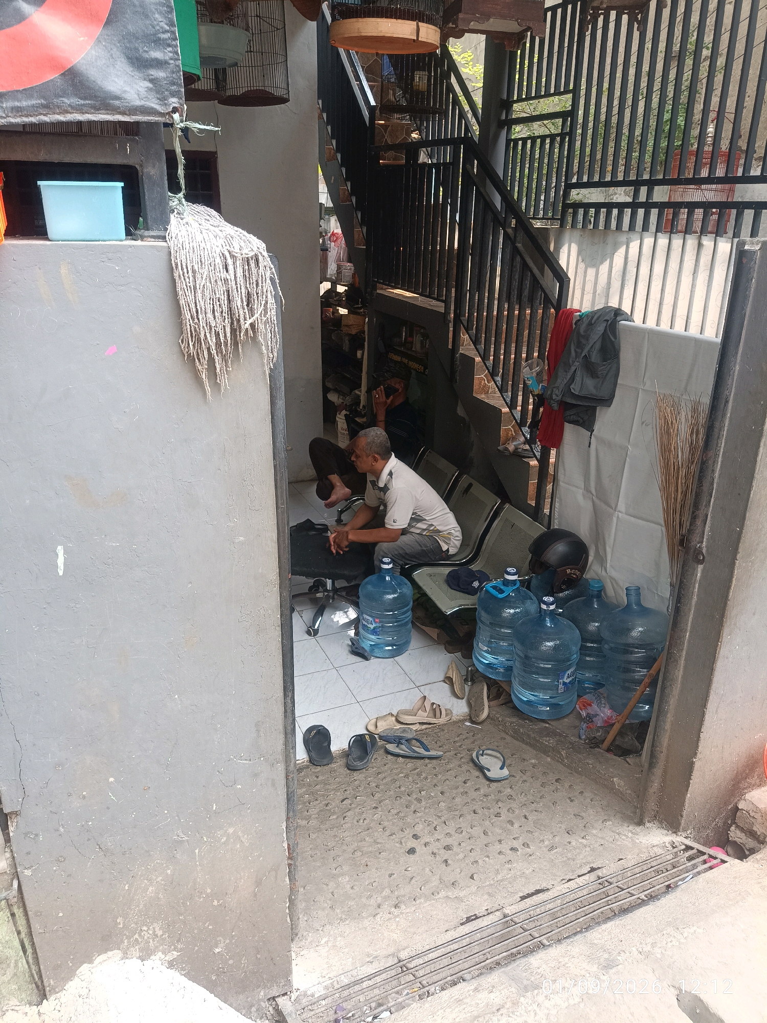 An outdoor, unpaved area shows a man sitting on a rolling chair with a water cooler in front of him. He is wearing a white polo shirt and dark pants. Behind him, several large jugs of water are stacked. A set of stairs with a metal railing is visible in the background. The scene is lit by natural daylight.