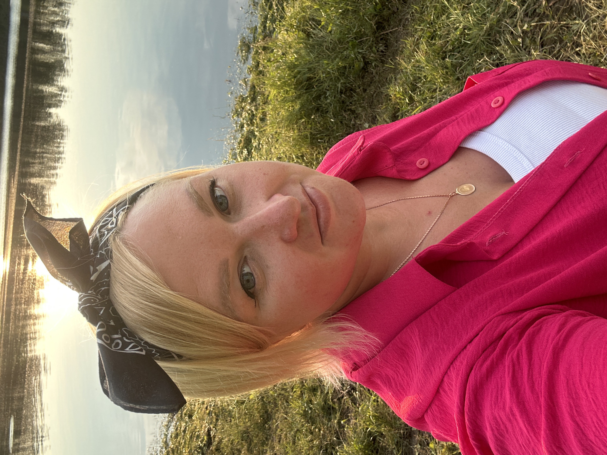 A medium shot, taken outdoors at dusk, shows a blonde woman with blue eyes wearing a bright pink collared shirt over a white tank top and a black bandana tied around her head. She is looking at the camera with a slight smile. The background consists of a body of water with the sun reflecting on its surface, and a grassy bank. The image appears to have been taken on the Kama River embankment in the city of Chaikovsky, Russia, given the characteristic sunset reflection on the water and the natural landscape.