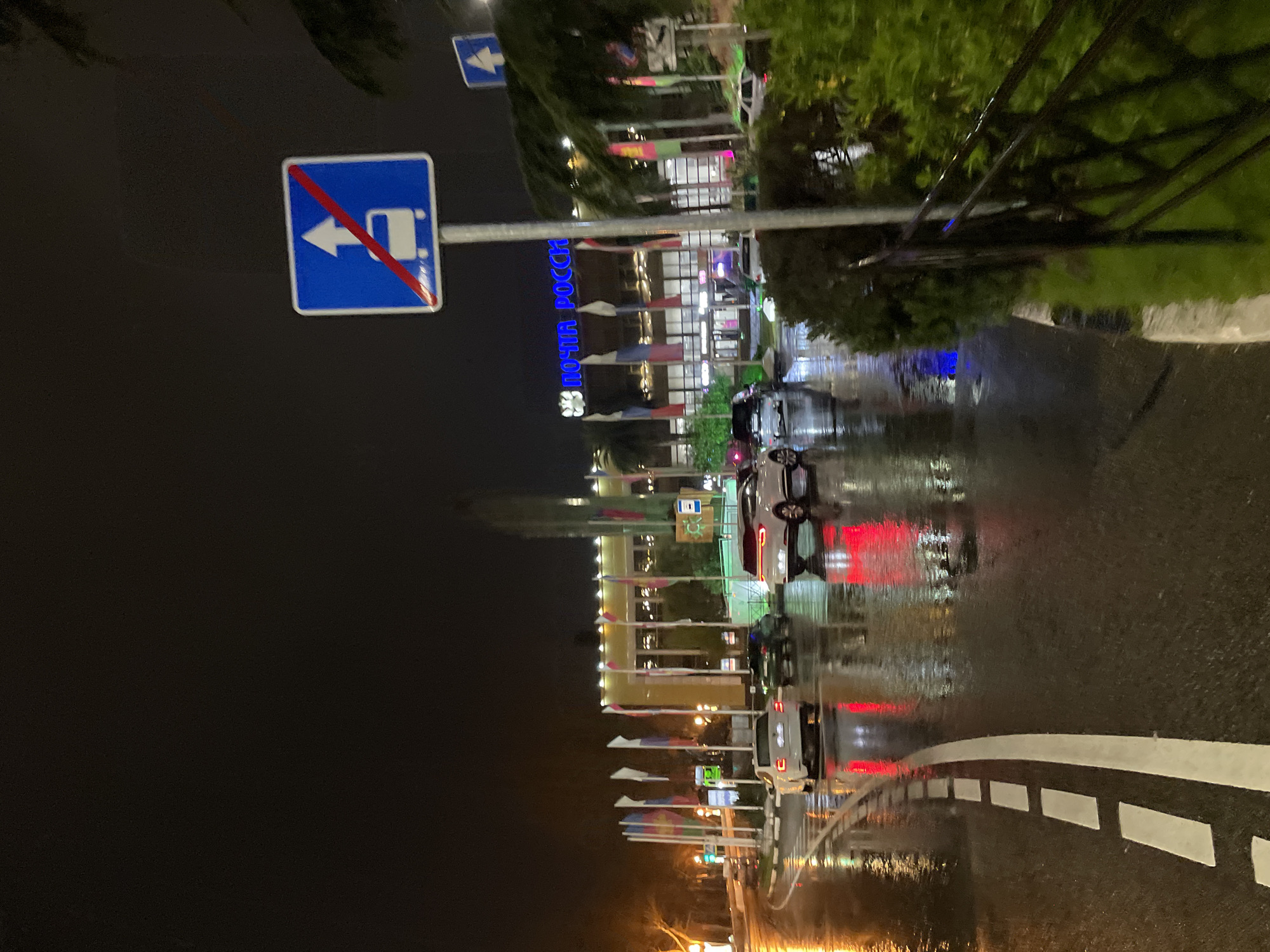 The image, taken at night in the city of Sochi, Russia, shows a wet road surface reflecting light from buildings and cars. A blue and white road sign indicates that turning left is prohibited for trucks. The Russian Post building ("ПОЧТА РОССИИ") is visible in the background, with multiple Russian flags displayed. Cars are parked along the road and driving on it, reflecting their taillights in the wet surface. The environment appears to be urban, with street lighting and trees present. The overall scene is characterized by reflections and the blurred effect of rain.