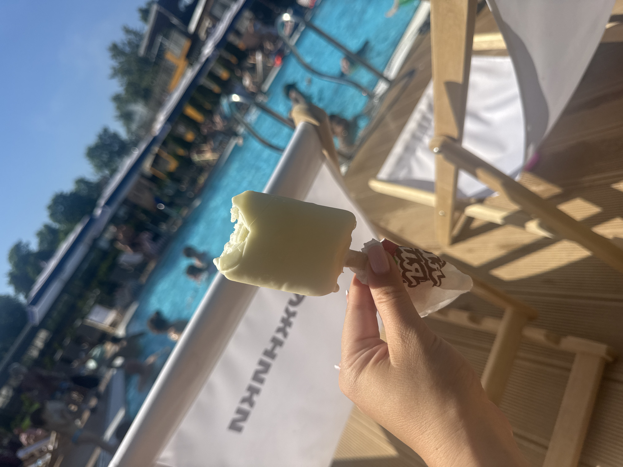 The image captures an outdoor recreational pool facility in Moscow, Russia, under a clear blue sky on a sunny day. In the foreground, a person's hand with light pink nail polish holds a white, rectangular popsicle, from which a bite has been taken, revealing the white interior. The popsicle stick is visible, and it is wrapped in a white paper wrapper featuring a red and dark brown/black stylized logo or pattern. The hand is positioned over a light-colored wooden surface, likely decking.
Behind the hand, a white fabric surface, possibly a lounge chair backrest, displays large, dark grey Cyrillic characters: "НККНЖ" (transliterated as NKKHZh). A light-colored rail or beam runs horizontally across the midground, partially obscuring the view of the pool.
The background reveals a vibrant blue swimming pool filled with numerous people swimming, wading, or standing. Metal ladders are visible, providing access to and from the pool. Beyond the pool, various blurred structures, possibly concession stands, changing rooms, or covered seating areas with awnings, are present, along with lush green trees, indicating a natural environment surrounding the facility. The scene depicts a lively leisure atmosphere.
