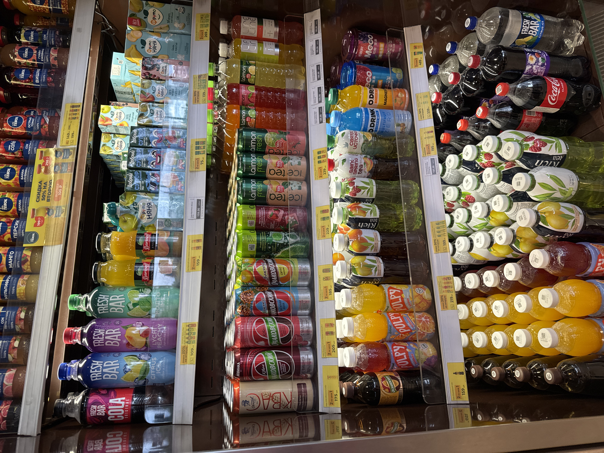 The image shows a refrigerated display case in a grocery store. The shelves are stocked with a variety of beverages, including sodas, juices, and teas, in bottles and cans. Many of the products are branded with Russian labels. A price tag is visible for "Ochakovskiy kvass" at 205 rubles. Another price tag indicates a sale of "fresh bar citrus ice" at 159 rubles. The overall environment suggests a retail setting focused on chilled drinks. The scene does not contain any people.