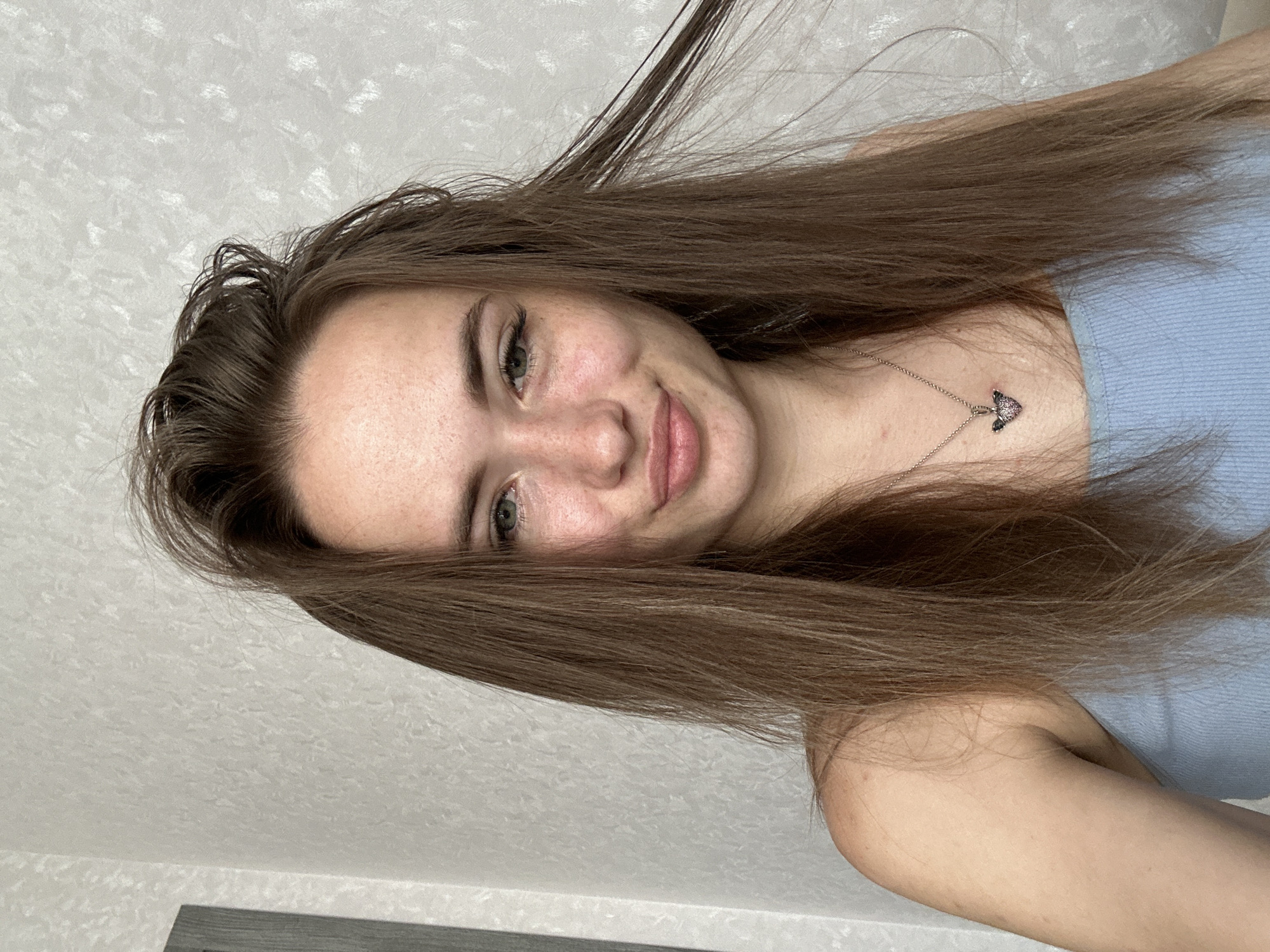 An unidentifiable young woman with long, brown, slightly disheveled hair, light skin with freckles, and green eyes is pictured from the chest up against a textured, light-colored wall. She is wearing a light blue tank top and a silver necklace with a heart-shaped pendant. She is looking directly at the camera with a slight smirk and her mouth puckered. The lighting suggests an indoor setting, possibly in a room with natural light coming from the side. The background wall has a subtle pattern, and to the left of the woman is a dark vertical object, possibly a doorframe or furniture. The overall impression is a casual, self-taken photograph. There is no information about the location in the city of Unknown, Unknown.