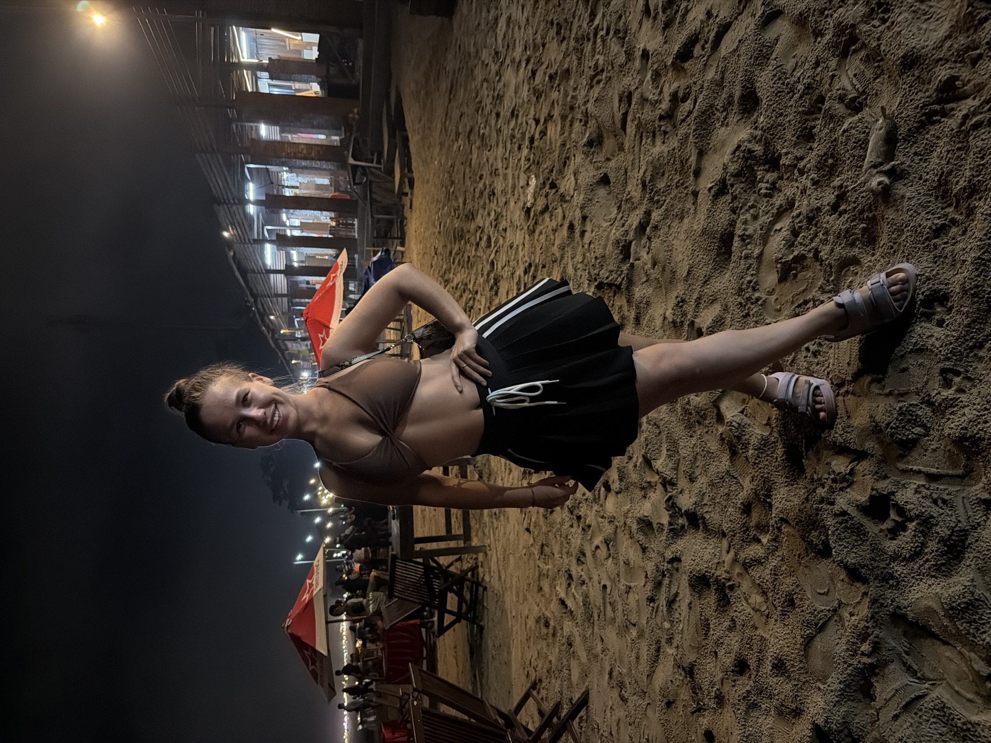 A young woman stands on a sandy beach at night, facing the camera with a smile. She is wearing a brown bikini top and a black pleated skirt with white stripes. Her hair is pulled up in a bun. She is wearing sandals. The background shows a dimly lit beach scene with tables, chairs, and red umbrellas, suggesting a beachfront restaurant or bar. Lights from buildings and string lights are visible in the distance. The sand is textured with footprints. The scene is located in Kuta Selatan, Indonesia.