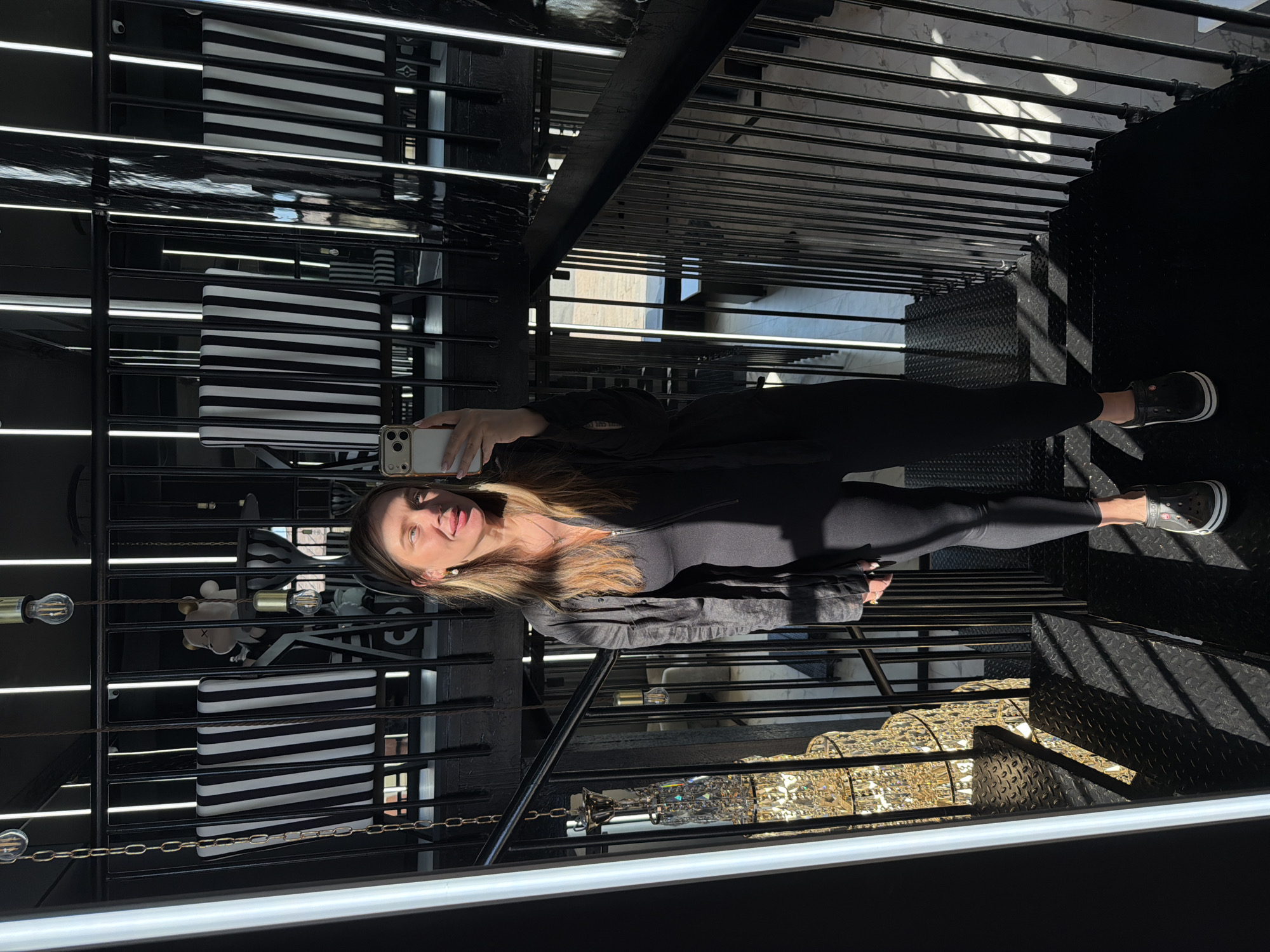 A medium shot captures a woman from the waist up, standing on a black metal staircase and holding her phone to take a selfie. She is wearing a black, tight-fitting jumpsuit with a zippered neckline and a black, unbuttoned overshirt. The woman has long, blonde-brown hair and is sticking her tongue out playfully. Her earrings are small and white, and she is wearing a necklace with a small pendant. Her feet are adorned with black Crocs clogs.
The background is dominated by a dark, industrial staircase with black metal railings. Sunlight streams through the railings, casting striped shadows on the steps. In the mid-ground, there are two chairs with black and white striped upholstery, and behind them, a large, elaborate crystal chandelier hangs from the ceiling. The overall aesthetic is modern and slightly edgy, with a mix of hard lines and glittering elements. The location appears to be indoors, possibly a stylish apartment or a commercial space.