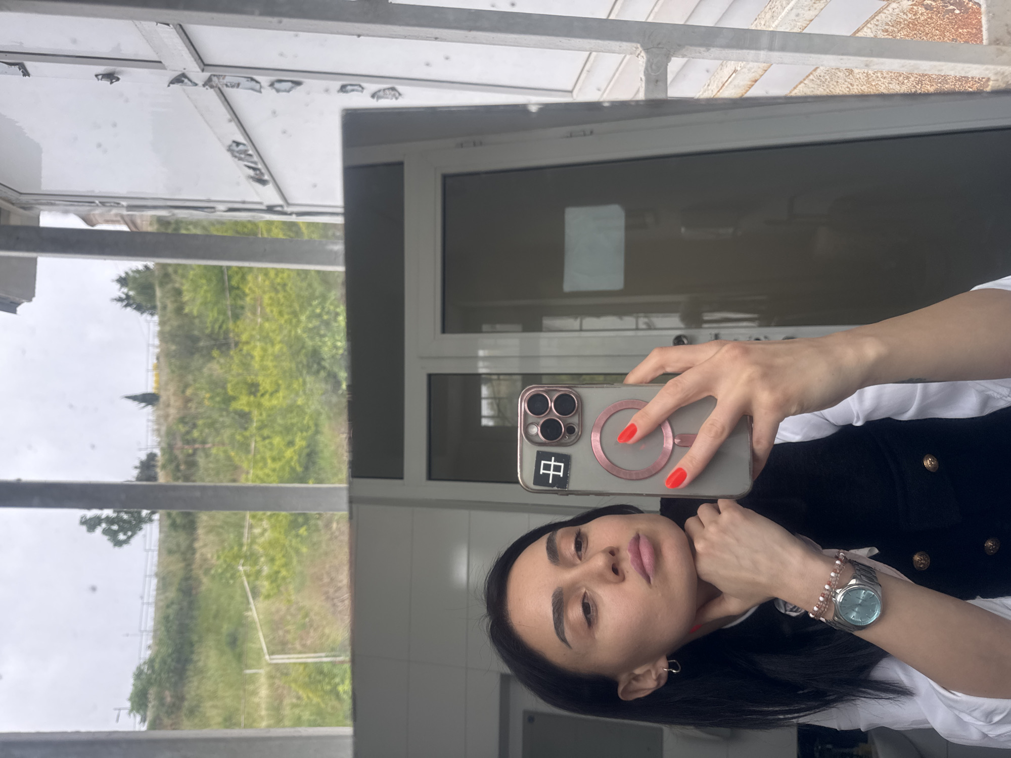 A young woman is depicted taking a mirror selfie indoors. She holds a smartphone, likely an iPhone with a transparent case and a rose gold MagSafe ring, in her right hand. Her fingernails are painted a bright red. On the phone case, a black square sticker with the white Chinese character "中" (zhōng) is visible.
The woman has dark hair and a neutral expression, looking directly at the phone's camera. She wears a white collared shirt under a black vest with gold buttons. On her left wrist, she has a silver watch with a light blue face and a multi-strand bracelet made of pink and white beads. A small silver hoop earring is visible in her left ear.
The indoor setting appears to be a functional space, possibly a restroom or hallway, with a large, clean mirror. The reflection in the mirror reveals an outdoor scene: a green, grassy landscape with trees under what appears to be an overcast sky. Above the landscape in the reflection, there's a light-colored, possibly metallic or concrete, architectural structure, suggesting a balcony, roof overhang, or similar outdoor building element. The lighting indicates it is daytime. The image offers a glimpse of both the woman's personal moment and the external environment, providing context within Tbilisi, Georgia.