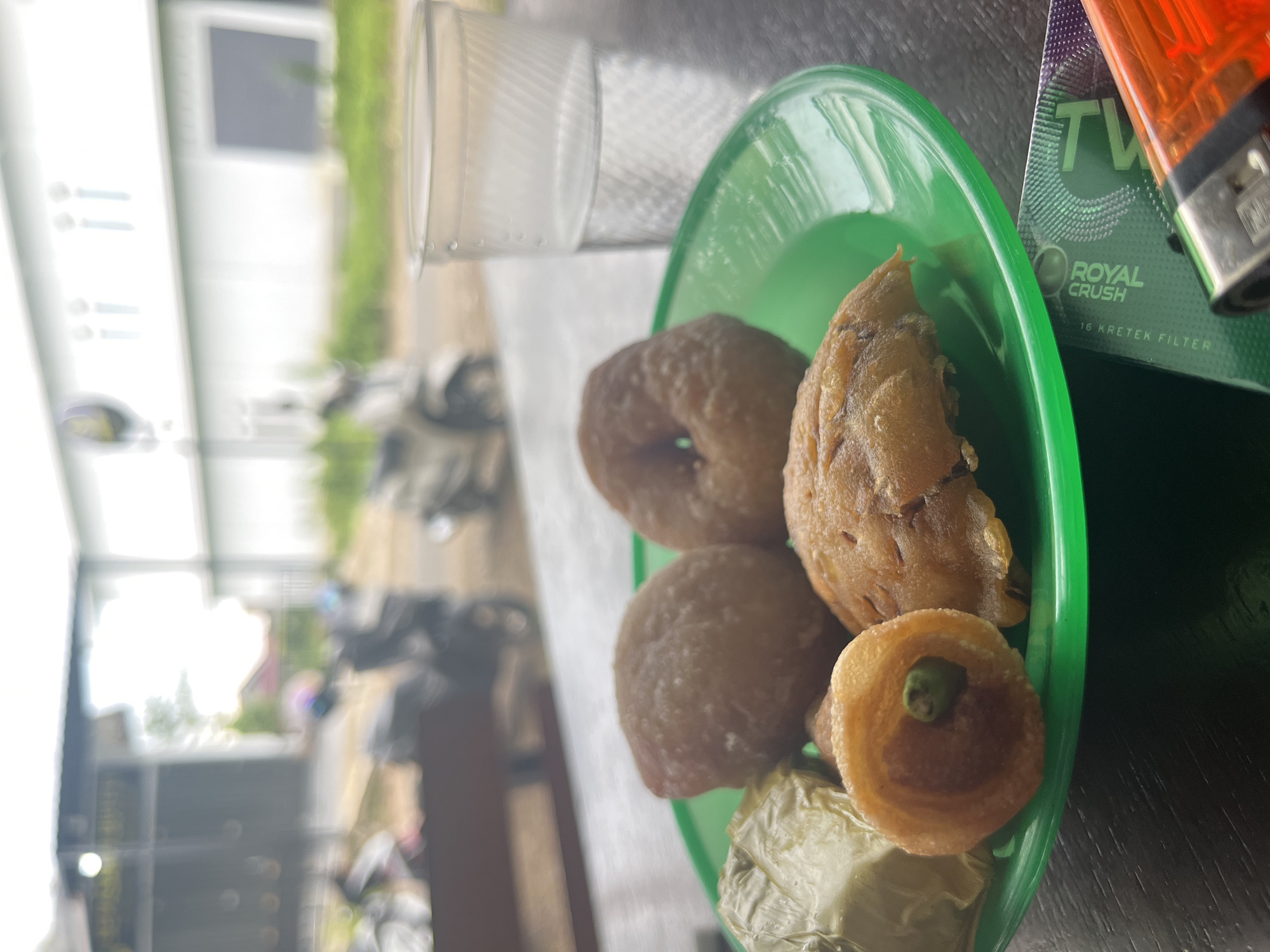 An outdoor scene in Keudematangkuli, Indonesia, features a green plate filled with fried snacks, including what appear to be donuts and some wrapped items. A clear plastic cup of water sits to the right of the plate, and a lighter and cigarette pack are visible in the foreground. In the blurred background, there are buildings, fences, and parked motorcycles, suggesting a roadside eatery or a casual public space. The lighting indicates it is daytime, possibly late afternoon or early morning.
