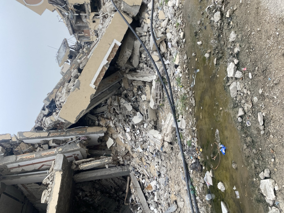 The image displays a scene of widespread destruction, likely the aftermath of a significant event such as an earthquake, bombing, or demolition. The primary focus is a severely damaged multi-story building, its structural integrity completely compromised, resulting in a large pile of rubble and debris.
The collapsed building dominates the left and central portions of the frame. Concrete slabs, which once formed floors and walls, are fractured, tilted at various precarious angles, and piled haphazardly. Twisted rebar protrudes from the broken concrete, indicating the reinforced nature of the original structure. Some sections of what appear to be exterior walls or balcony edges still show remnants of decorative patterns, possibly tiles or painted designs, suggesting the building once had an architectural finish. The upper parts of the structure, visible against a light, overcast sky, show more fragmented concrete and exposed interior elements.
To the right of the extensive rubble, the ground transitions to an unpaved area, a mix of dirt and scattered smaller pieces of concrete and general debris. A muddy, greenish-brown puddle or trench runs horizontally across this ground section, suggesting either stagnant water from recent rainfall or a broken utility line. Several dark, thick cables or hoses run across the middle of the image, stretching over the debris field and alongside the muddy water, possibly belonging to the building's infrastructure or emergency equipment.
Scattered amongst the dirt and smaller debris on the right side are various pieces of trash, including what looks like plastic packaging, a blue bag, and other discarded items. The overall impression is one of desolation and ruin. The sky above is uniformly light grey or white, indicating an overcast day with diffused lighting, offering no strong shadows. There are no people visible in the image, emphasizing the silence and emptiness of the devastated site.