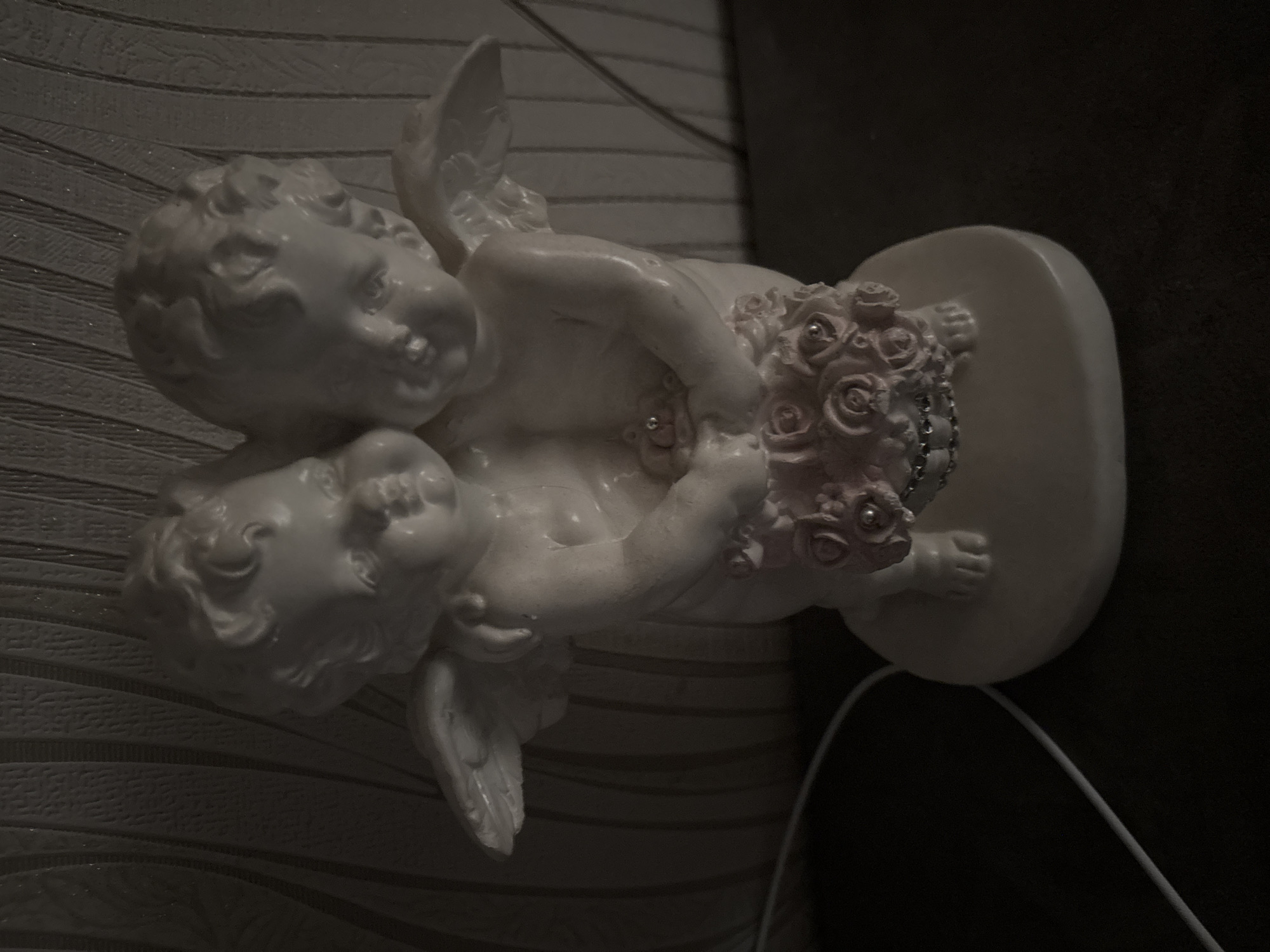 A white ceramic figurine depicting two conjoined cherubic figures stands on a dark, flat surface. Each figure exhibits curly hair, plump facial features, and small wings extending from their backs. They jointly hold a bouquet of pink roses, accented with silver beads, positioned centrally at their midsections. A singular silver embellishment is present on the upper torso of one figure. A thin white cable is visible traversing the lower left foreground. The background consists of a dimly lit wall featuring a vertical, textured stripe pattern. The image is underexposed, with soft shadows indicating low light conditions. The geographical location in Chernogorsk, Russia, cannot be ascertained from the visual content.
