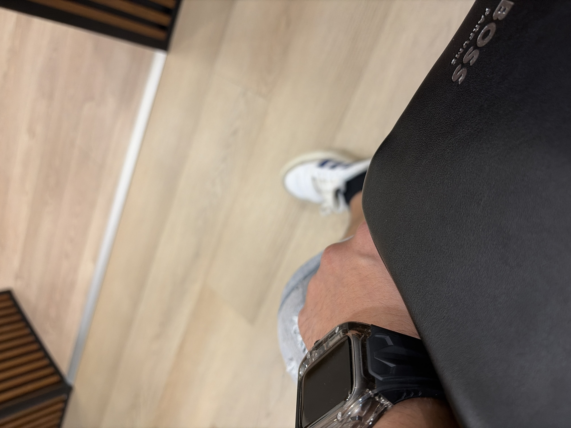 A close-up, angled shot from above captures a partial view of a person indoors. The subject wears a smart watch with a clear protective case on their left wrist and a white sneaker with blue stripes (likely Adidas) on their foot. They are holding a black, textured object, possibly a bag or portfolio, which prominently displays the silver text "BOSS PARFUMS" along its side. The background features a light wood-patterned floor, with a dark brown slatted structure visible in the top left and bottom left corners, and a silver metallic strip running along the floor in the mid-ground. The setting appears to be a modern indoor space, possibly a retail environment or a home. The time of day and any specific emotions are not discernible.