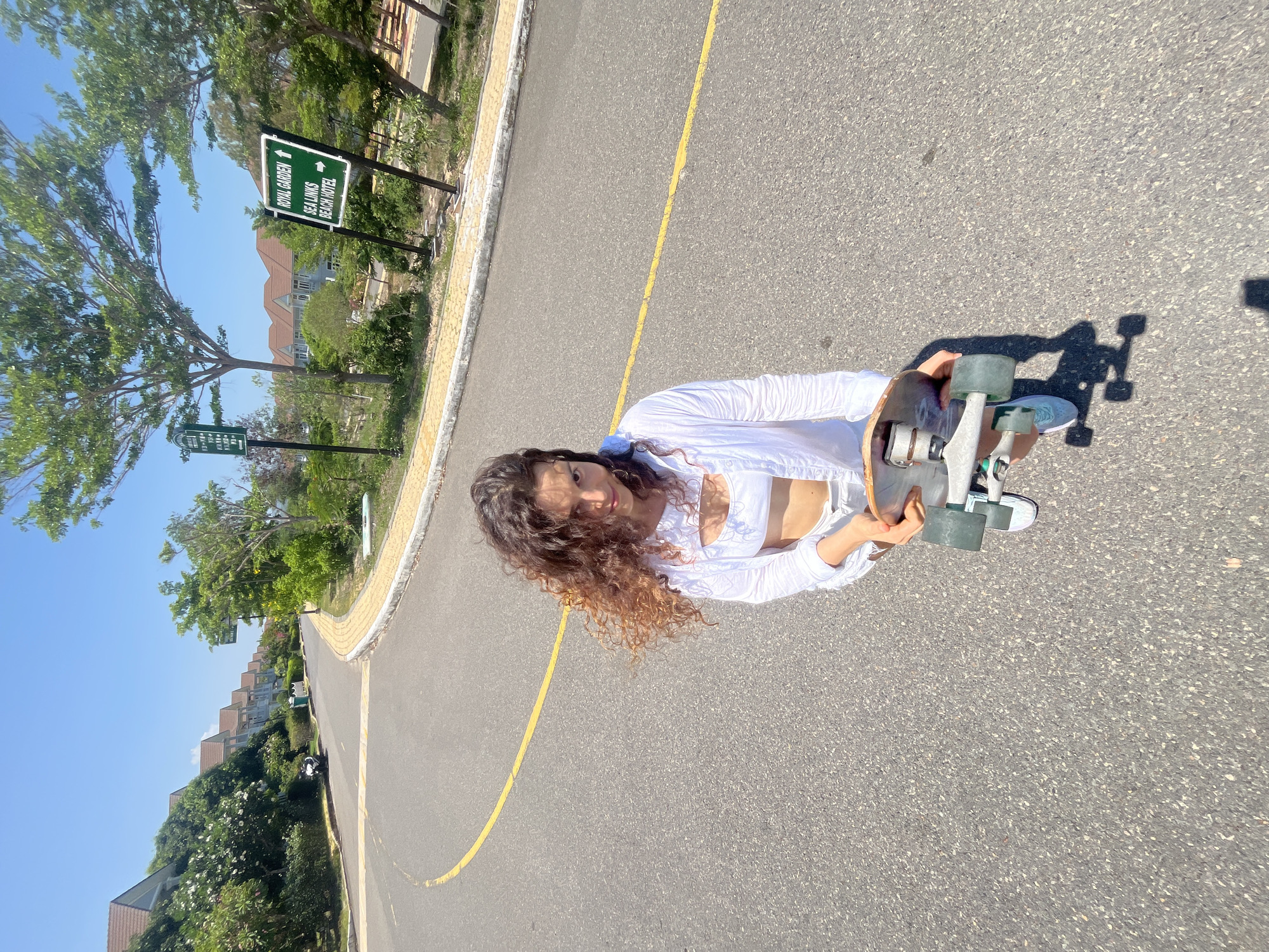 A young woman with curly brown hair is standing on an asphalt road in Phan Thiết, Vietnam. She is wearing a white open-front shirt, a white crop top, and white shorts. She is holding a skateboard in her hands, and it is positioned at her right hip. Her shadow is cast on the road to her right. The road curves to the left and has a yellow line painted down its center. To the left of the road, there are several multi-story houses with light-colored walls and brown roofs, surrounded by lush green trees and bushes. To the right of the road, there are more trees and two green directional signs. One sign points to "ROYAL GARDEN" and "SEA LINKS BEACH HOTEL" with arrows. The sky is clear and blue.