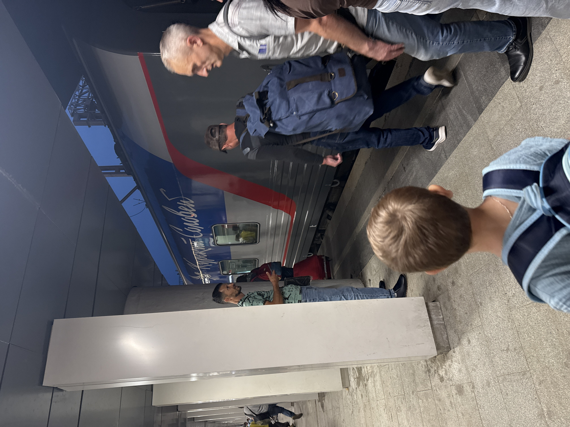 The image shows a train platform at a Moscow railway station, likely during the evening. A train with blue and red livery is stopped at the platform. Several people are present, some appearing to board or disembark. In the foreground, a young boy with a blue backpack is looking towards the train. To the left, a man with a beard in a green t-shirt and jeans stands near a white column. Further down the platform, a man wearing a dark shirt and baseball cap is walking towards the train, carrying a large blue backpack. To the right, a man with short gray hair in a checkered shirt and jeans is walking away from the train. The platform is paved with light-colored tiles, and white architectural columns line the station. The sky is visible above the train, suggesting an outdoor setting.