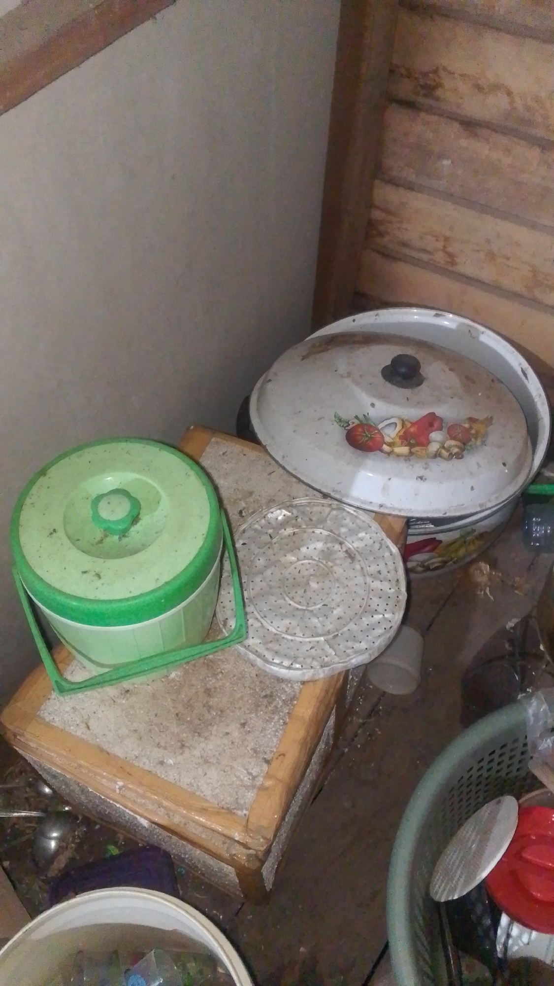 The image depicts a cluttered interior space, possibly a storage area or a room in a house located in Olak-Olak Kubu, Indonesia. In the foreground, on a wooden structure, sits a green plastic bucket with a handle. To the right of the bucket is a white circular strainer with numerous small holes. Further to the right, a large, white, lidded pot with a decorative image of vegetables is visible. The lid has a black knob. The floor appears to be made of wood and is covered with various items, including what looks like a green plastic basket containing more objects, one of which is a red lid. A light-colored wall is visible on the left, and wooden planks form a wall or partition on the right. The overall impression is one of disarray and storage of household items.