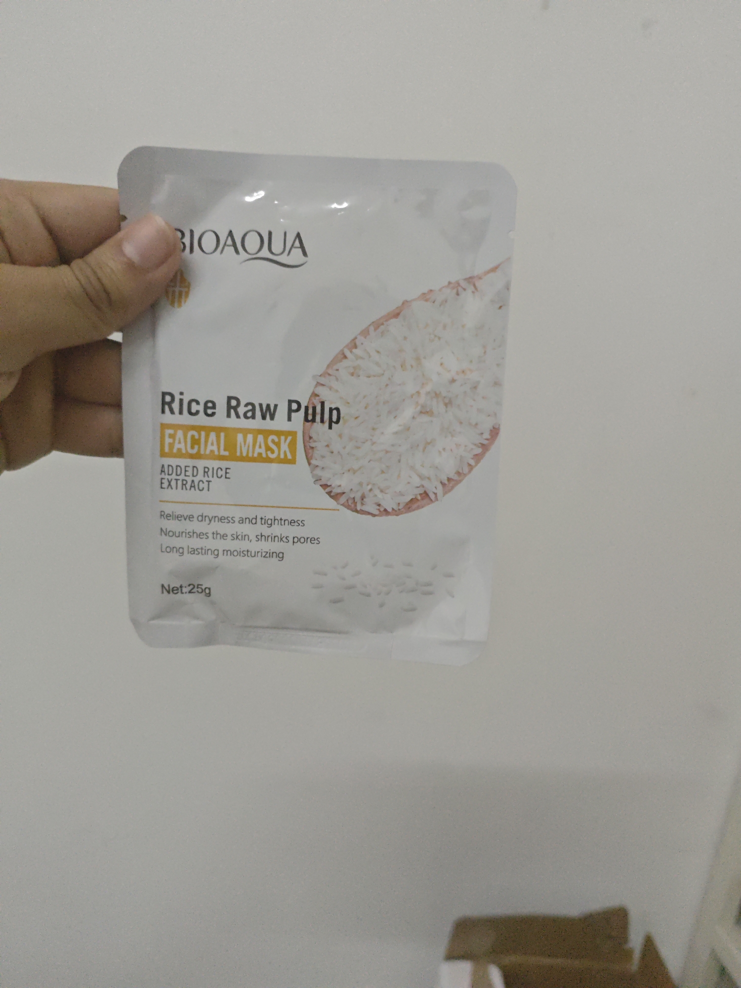 A hand holds a white facial mask package with a picture of rice on it. The package has "BIOAQUA" printed in black text at the top. Below that, it says "Rice Raw Pulp FACIAL MASK". Further down, it reads "ADDED RICE EXTRACT" and lists benefits: "Relieve dryness and tightness", "Nourishes the skin, shrinks pores", and "Long lasting moisturizing". The net weight is stated as "Net: 25g". The background is a plain white wall. This image was not taken in Chongqing High-tech Zone, China.