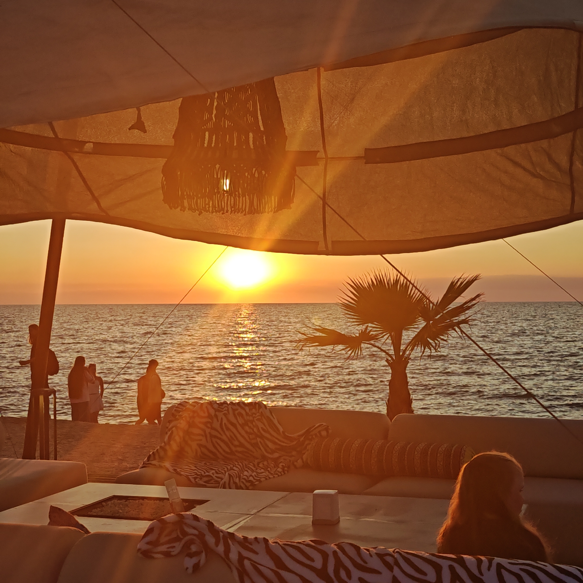 This is a medium shot, eye-level photograph of a beach scene at sunset. The foreground is dominated by a covered outdoor seating area with a white tarp overhead, illuminated by the warm, orange light of the setting sun. A decorative, macrame-style lampshade hangs from the tarp. A large, light-colored sofa with zebra-print blankets and patterned pillows is visible, with a low table in front of it. A small, white, square object sits on the table. To the right, a young person with long, blonde hair is partially visible, sitting on the sofa.
In the middle ground, a sandy beach meets the ocean. Several people are standing on the beach, silhouetted against the bright sunset. Some are facing the water, while others appear to be conversing. A small palm tree is situated on the beach.
The background features the ocean, with gentle waves reflecting the golden light of the sunset. The sun is a bright, intense orb low on the horizon, casting a shimmering path across the water. The sky above is a gradient of orange and pale yellow.