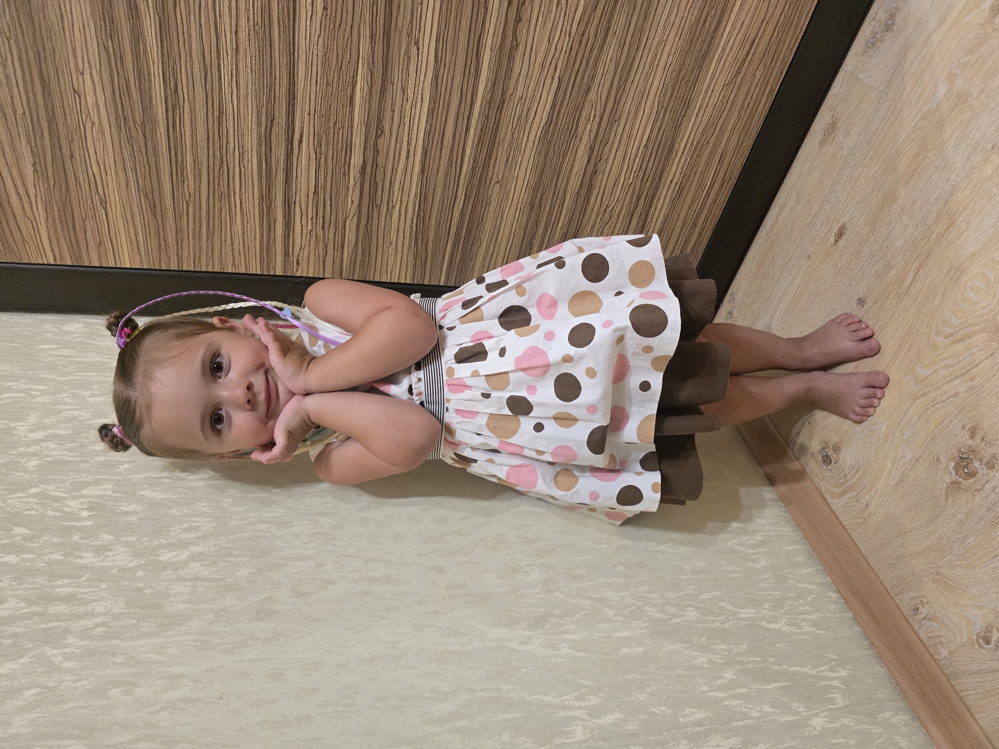 A young Caucasian girl with blonde hair styled in two buns with colorful strands is standing in a corner of a room, facing the camera. She is wearing a white dress with a pattern of pink, brown, and tan polka dots, and a brown underskirt. Her hands are placed on her cheeks, and she has a sweet expression on her face. The room has a textured beige wallpaper on one wall and a wooden door on the other. The floor is made of light-colored wood laminate. The lighting appears to be artificial and evenly distributed. The scene is indoors, likely a home environment, in an unspecified location.
