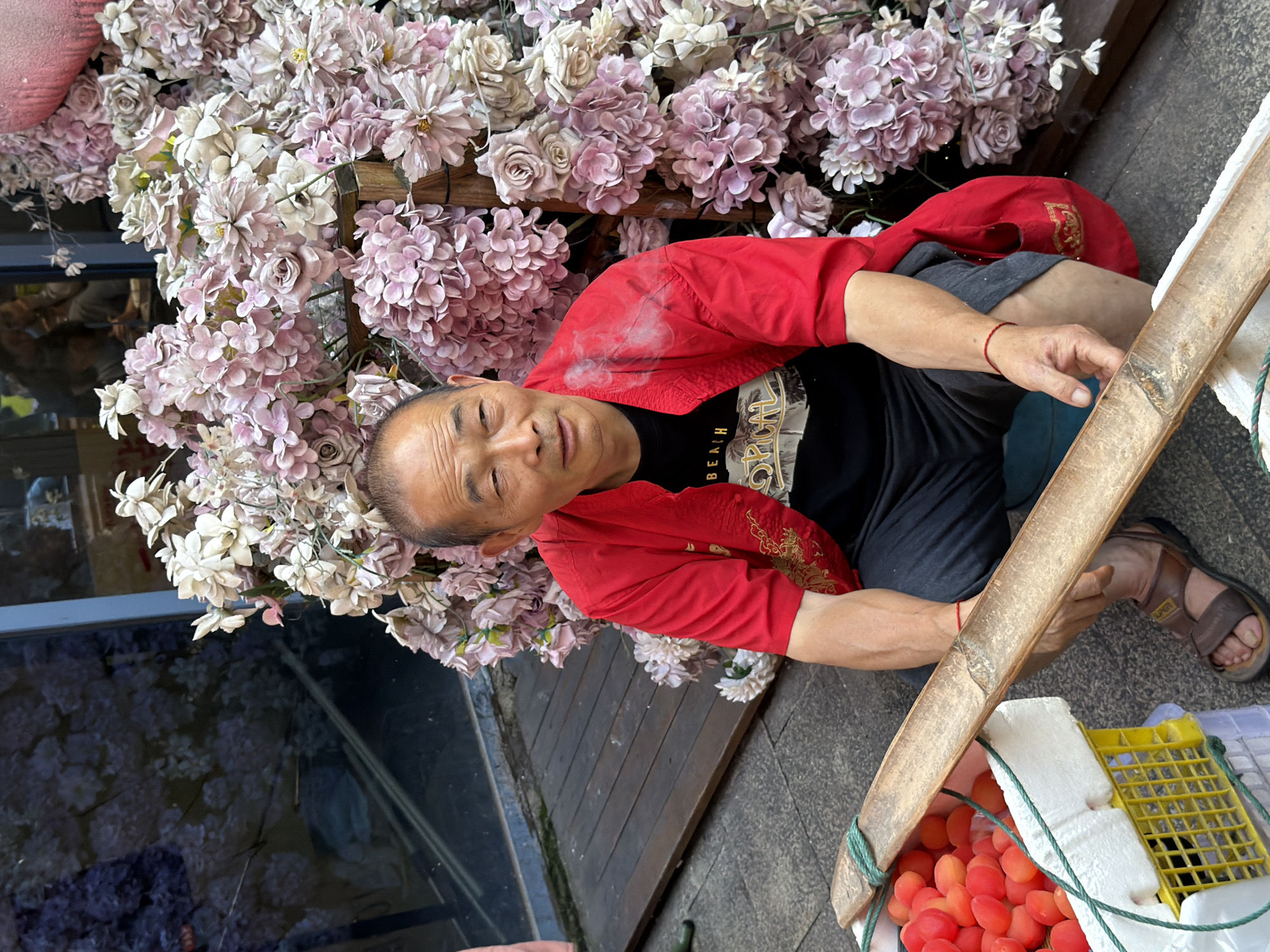 In Yuzhong District, China, an elderly East Asian man with short, dark hair and a weathered face sits on a wooden deck. He is wearing a bright red jacket over a black t-shirt with a graphic print and dark grey shorts. His right hand is resting on a makeshift ramp made of bamboo, which is positioned over a white styrofoam container filled with small, red, plum-like fruits. A yellow plastic crate is visible beneath the styrofoam. The man has a red string tied around his left wrist. Behind him, a large arrangement of artificial pink and white flowers, resembling roses and hydrangeas, is attached to a wooden structure. The background shows a building with large glass windows, reflecting blurry images of people and the surrounding area. The ground is paved with stone. The overall scene appears to be an outdoor marketplace or a street vendor setup.