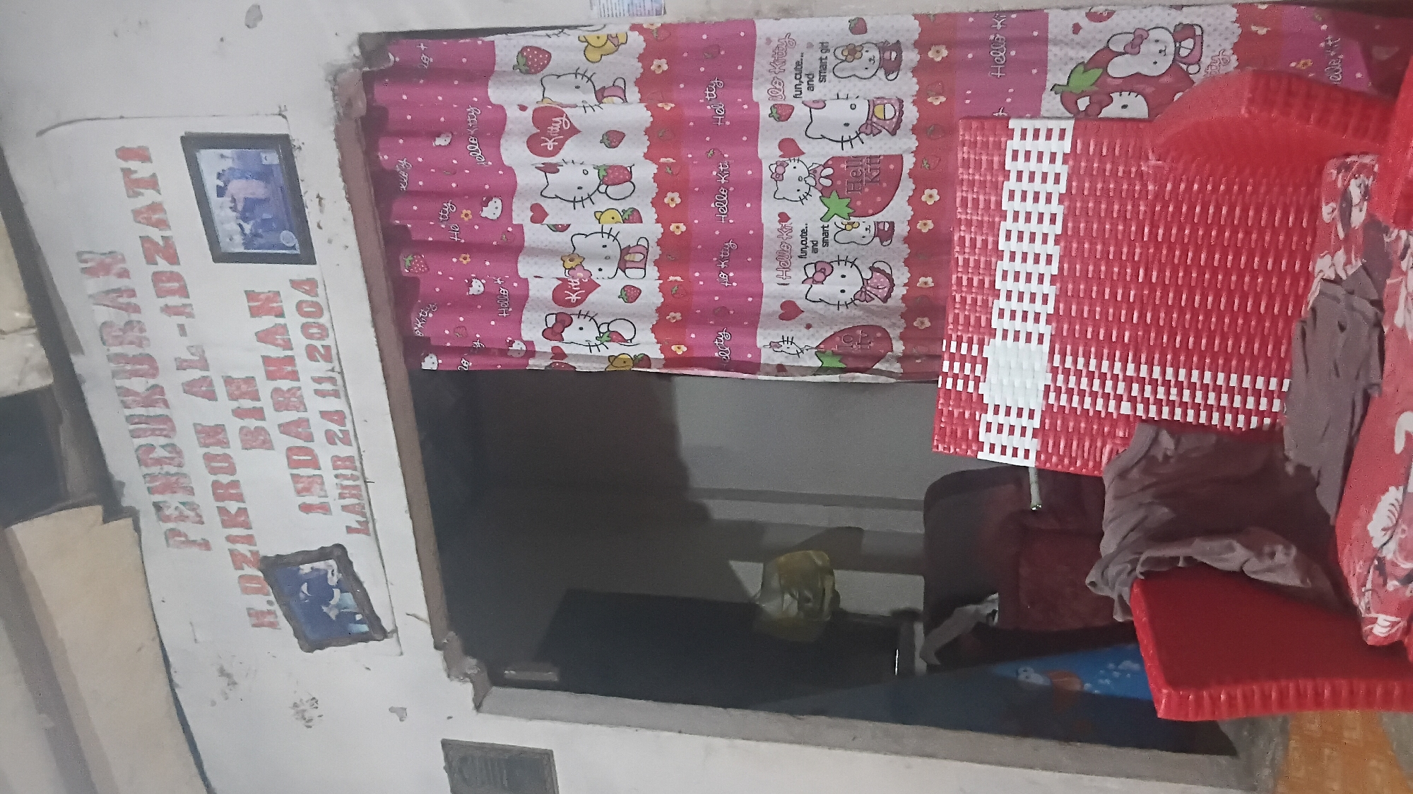The image depicts an indoor scene, likely a living room or a common area within a house. Above a doorway, a white sign with red lettering reads "PENSUKUBAN H. DZIKRON AL-IDZATI BIN INDARHAN LAHIR 24.11.2004." This text appears to be a memorial or biographical inscription. Two framed photographs are affixed to the wall above the sign. One shows a group of people, possibly at an event, and the other contains a picture of what appears to be a religious ceremony.
In front of the doorway, a pink curtain with a Hello Kitty pattern hangs. To the right of the doorway, there is a red wicker chair with a white and red woven pattern on its back. Another red armchair is visible to the left, partially obscured by a grey cloth. The environment suggests a modest, perhaps older, dwelling. While the specific location within Prabumulih, Indonesia, cannot be determined from the image alone, the presence of Indonesian script on the sign strongly indicates it is located within Indonesia. The overall impression is one of a personal and perhaps commemorative space within a home.