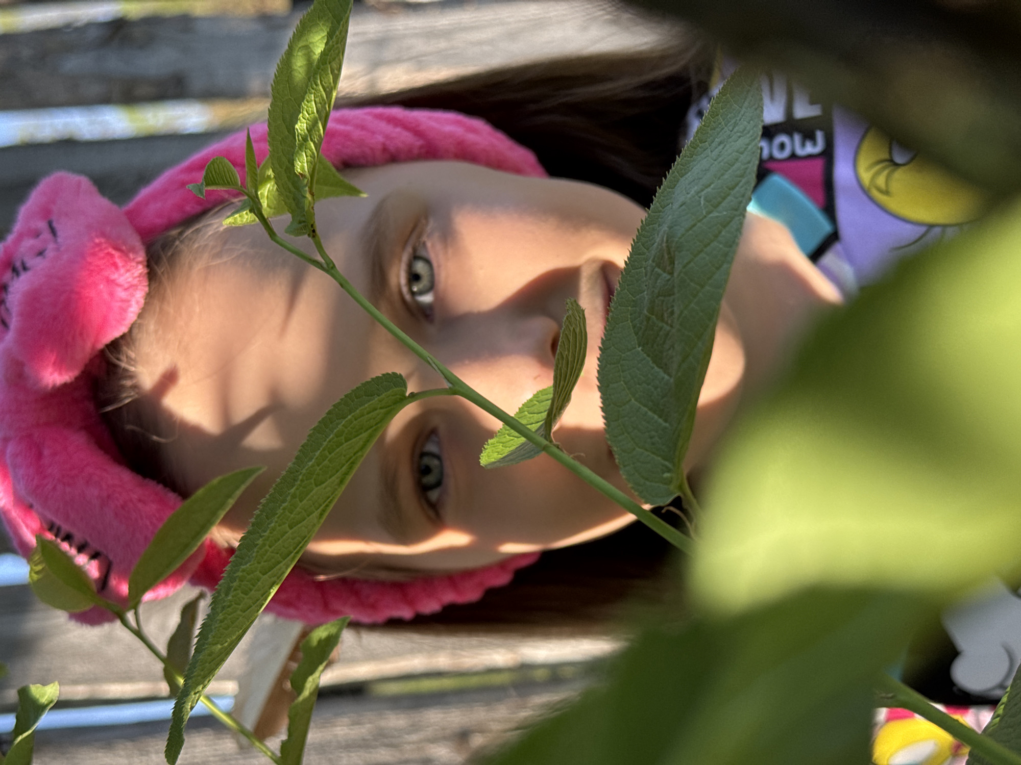 A young girl is seen in a partial view behind green leaves. She has light skin, green eyes, and brown hair. She is wearing a pink plush headband with what appears to be bunny ears. Her clothing is a purple shirt with cartoon images, including a yellow character. The background includes a gray, possibly wooden, surface. The location is Rusalevka, Russia.