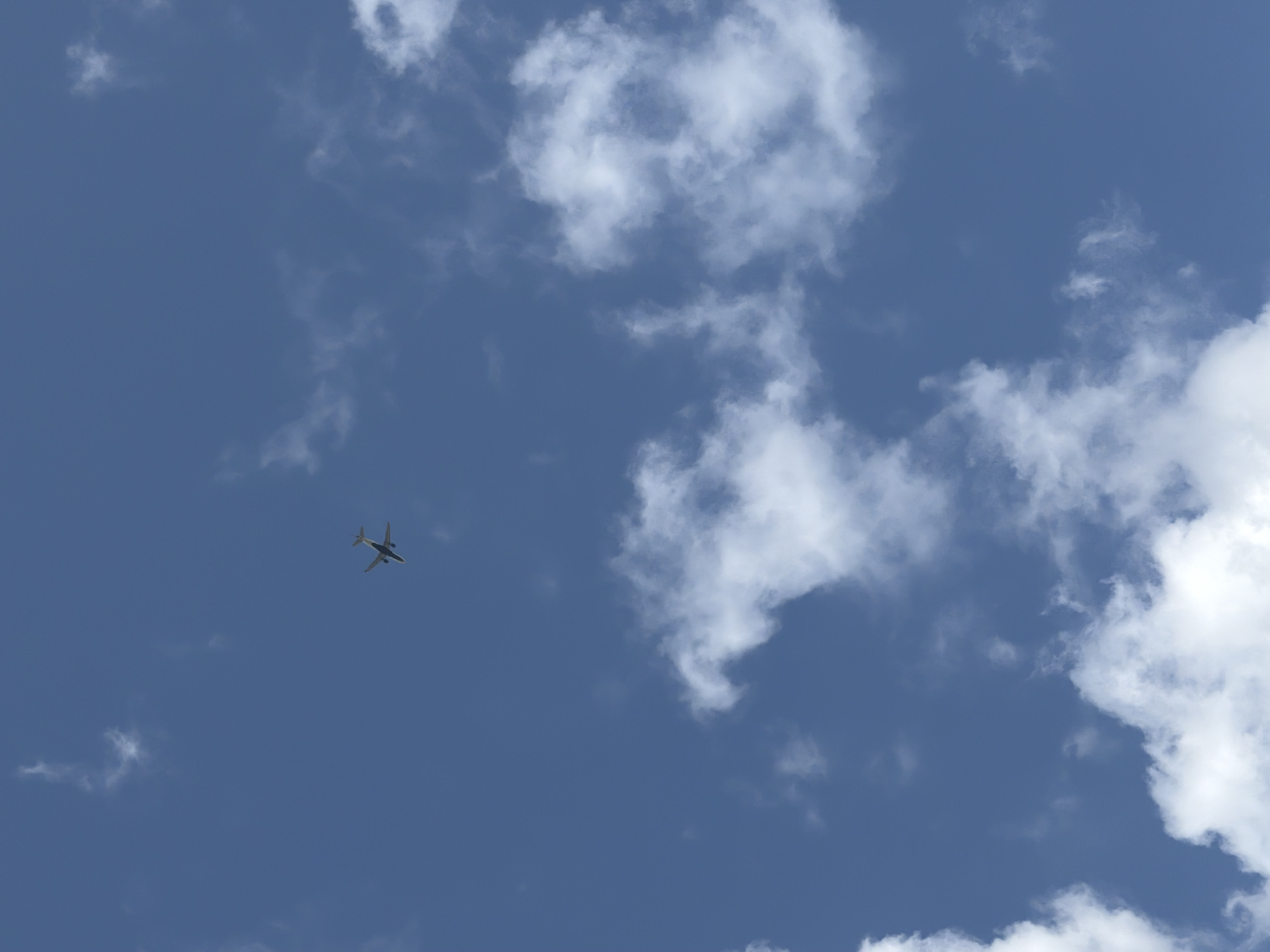 A small, dark-colored airplane is captured in mid-flight against a vast, blue sky dotted with fluffy white clouds. The airplane is positioned in the upper left quadrant of the image, appearing as a silhouette with visible wings and tail. The vast expanse of the sky suggests an aerial perspective, likely from the ground looking upwards. The image does not contain any discernible landmarks or features that would allow for its identification within the city of Pittsburgh, United States of America. The scene is natural and unobstructed, with no people or immediate interactions visible. The environment is exclusively the sky and clouds.