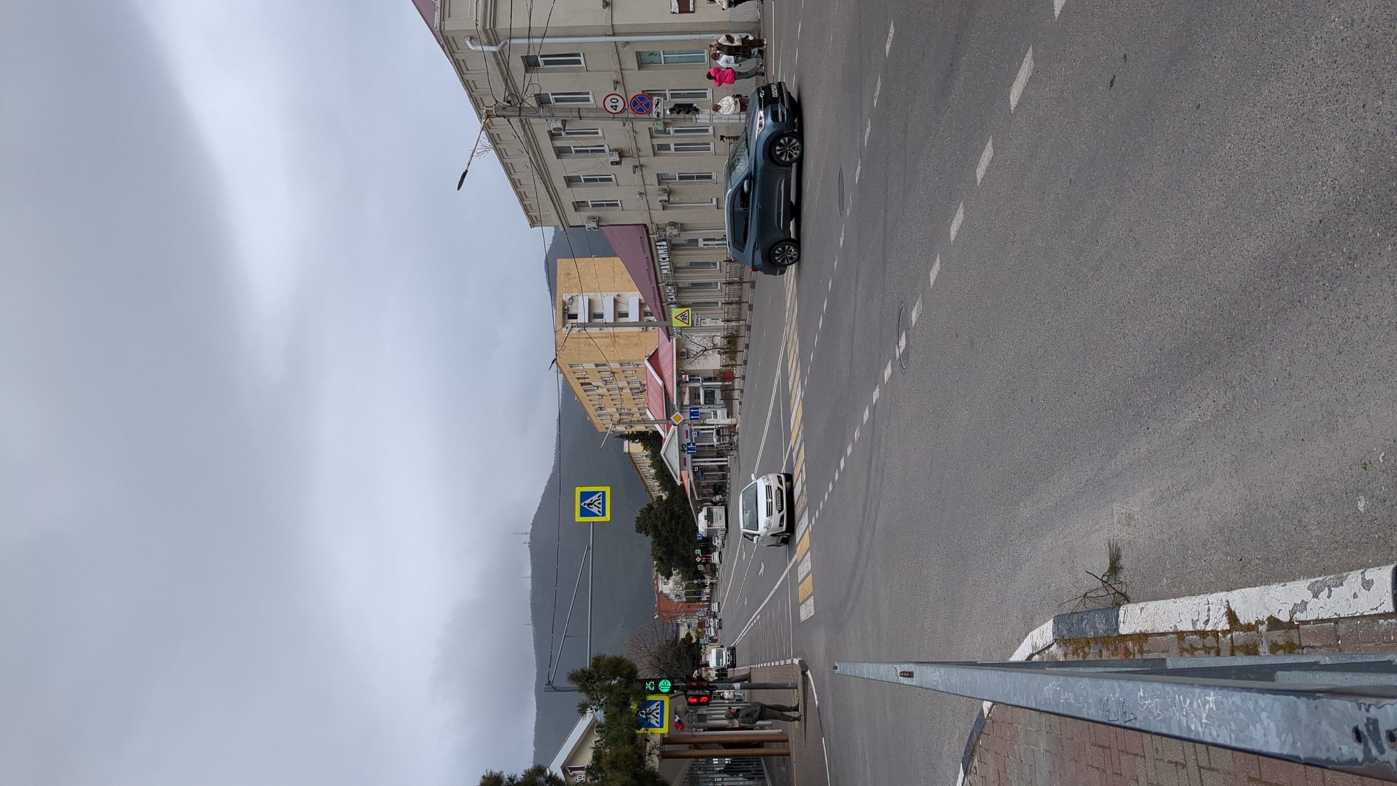 The image depicts a street in Gelendzhik, Russia, on an overcast day. The street slopes downhill, with buildings on the right and a mountain in the background to the left. A dark blue car is parked on the right side of the street, while a white car drives down the road. Pedestrian crosswalks with corresponding signs are visible. There are people in front of the buildings on the right. A traffic light is on green. A fence and sidewalk are in the foreground.