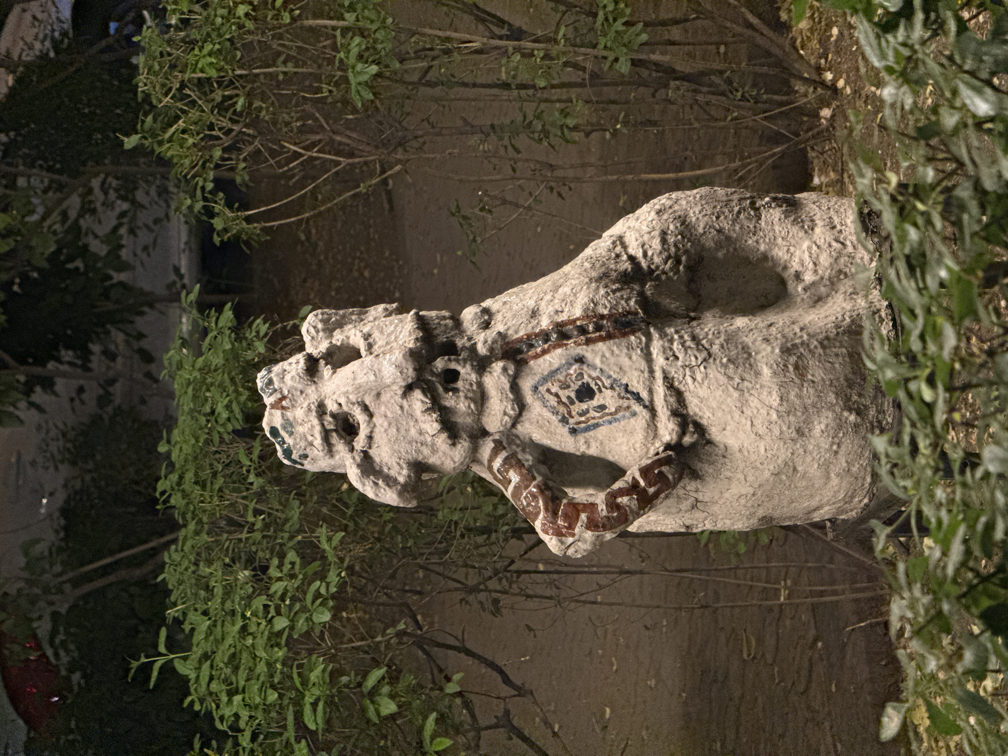 Here's a description of the image:
The image shows a stone horse sculpture, likely a garden ornament, set amongst greenery, probably bushes. The sculpture is heavily textured, with weathered and cracked surfaces, painted white. It has details such as a decorated harness or saddle blanket with geometric patterns in blue and brown. The sculpture is positioned near a stone path or low wall. The setting appears to be a park or garden, and the lighting suggests it is evening. The location is Yerevan, Armenia.