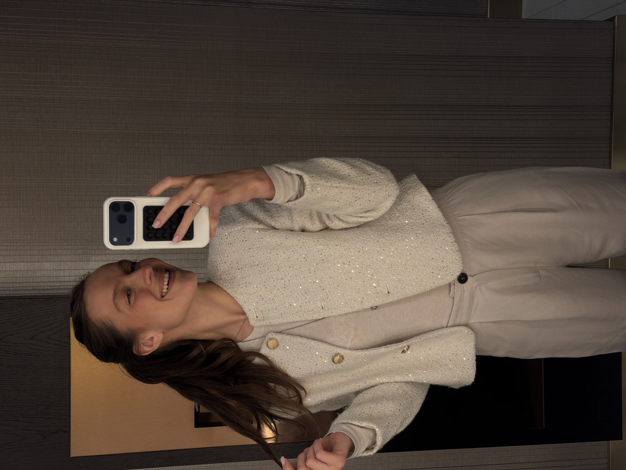 A young woman is captured in a mirror selfie, smiling broadly, appearing happy and cheerful. She is positioned indoors, likely in a hotel room or apartment in Budva, Montenegro, given the context. The image is taken from a slightly low angle, showing her torso and face.
She has long, dark brown hair and is wearing a stylish, textured off-white or light beige jacket adorned with subtle sequins and featuring gold-toned buttons. Underneath, she wears a plain light beige long-sleeved top and light beige pleated trousers. On her right hand, holding the smartphone, a silver ring is visible on her ring finger.
The woman is holding a white or light-colored smartphone with a prominent black camera module and a black textured grip on the back of the case. Her reflection is visible in what appears to be a mirror or a dark, reflective surface that forms part of the background. The background features a dark brown/grey textured wall with subtle horizontal lines, suggesting a modern interior design. The lighting is artificial and soft. There is no visible text in the image.