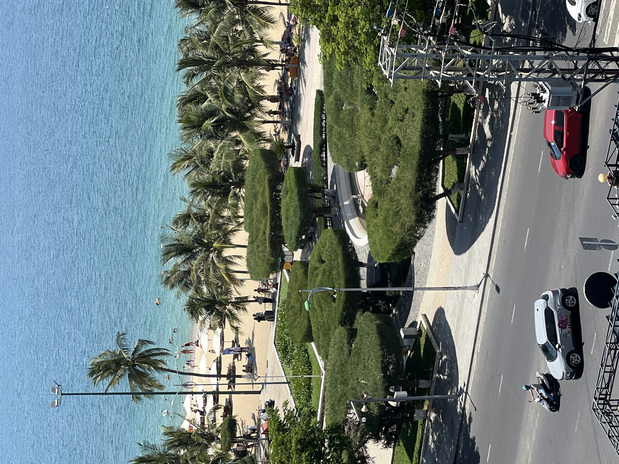 The image captures a panoramic view of Nha Trang, Vietnam, showcasing the city's vibrant beachside and urban landscape. The scene is divided into three distinct horizontal sections.
The uppermost section displays the cerulean waters of the sea, with gentle waves lapping at the shore. Several individuals are visible in the water, swimming and enjoying the ocean. The beach itself is a strip of golden sand, populated with sunbathers relaxing under numerous white umbrellas and beach chairs. Palm trees dot the shoreline, their fronds swaying in the breeze.
The middle section transitions to a manicured park area bordering the beach. This area features meticulously trimmed, rounded trees arranged in a decorative pattern, interspersed with lush green grass and smaller plants. Several pathways wind through the park, and at least one street lamp with multiple lights stands tall. A few individuals are seen walking or standing in this area, some with luggage, suggesting they might be tourists or hotel guests.
The lowest section of the image depicts a city street running parallel to the park and beach. A grey SUV with decorative stickers on its doors is parked or driving on the road, and a person is riding a scooter. Further down the street, a red car is in motion. Electrical infrastructure, including a utility pole with transformers and wires, is prominently visible on the right side of the road. The presence of a crosswalk indicates this is a pedestrian-accessible area. The overall impression is of a bustling coastal city on a sunny day.