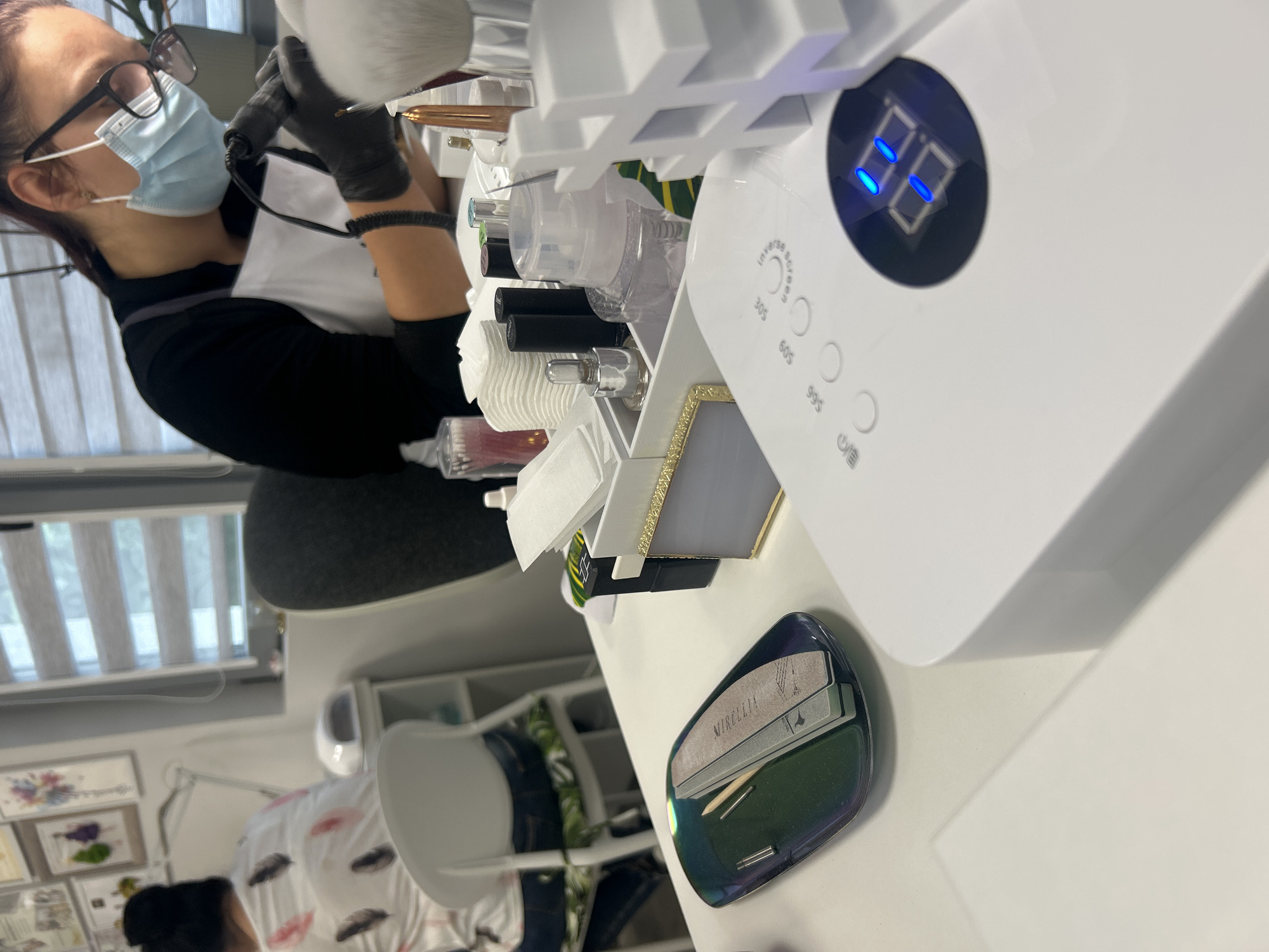 A sideways indoor shot shows a person wearing a light blue disposable face mask and black gloves is working on nails at a white manicure table. The person has on a black long-sleeved shirt and a white apron. The manicure table has a UV lamp with a digital display showing "88", and buttons labeled "30s", "60s", and "99s". There is a white organizer with various nail care products on the table, including bottles of nail polish, brushes, and cotton pads. A black and green nail palette with nail files and other tools on it is on the right. The background shows a window with blinds and a white chair. Artwork hangs on the wall in the bottom left. The location is likely a nail salon in Unknown, Unknown.