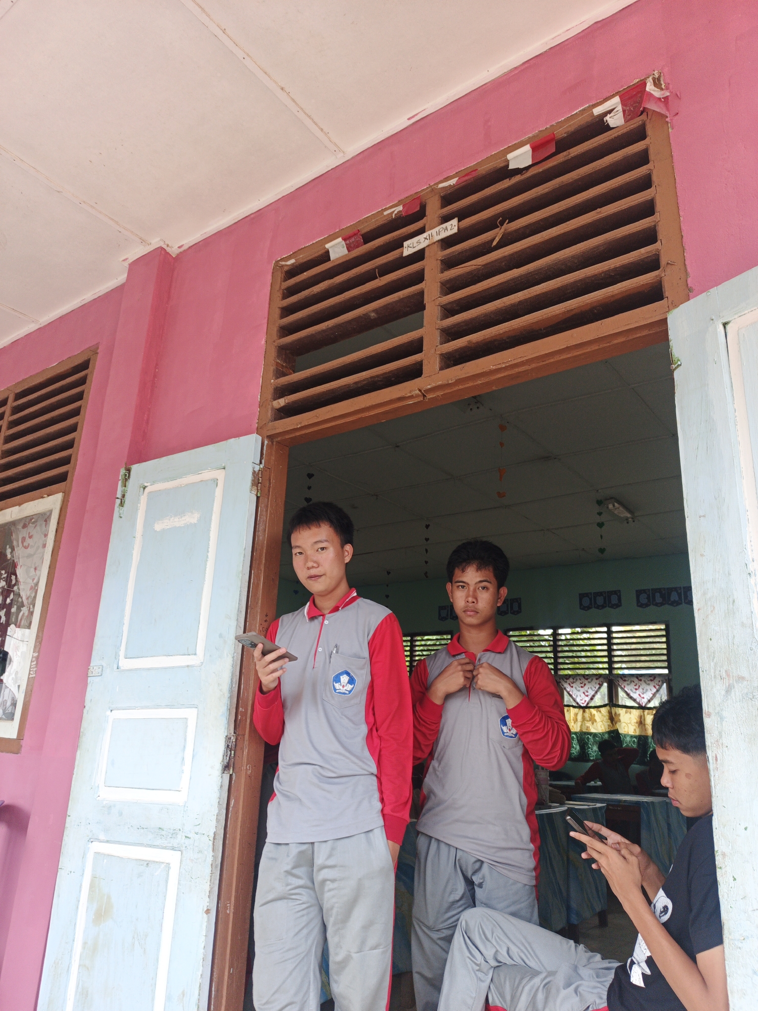 The image depicts a scene outdoors, likely in front of a classroom or school building in Tanjung Setia, Indonesia, given the visible signs and uniforms. Two young men, appearing to be students, are standing in a doorway. The one on the left, wearing a red and gray polo shirt and gray pants, holds a smartphone in his left hand. The young man to his right, similarly dressed in a red and gray polo shirt and gray pants, is adjusting the collar of his shirt. A third young man, partially visible on the far right, is seated and looking at his smartphone. The building features a pink exterior wall and a wooden-slatted window above the doorway. Inside, the classroom appears to have desks and chairs, with other individuals seated. The overall impression is a casual moment captured outside a school.