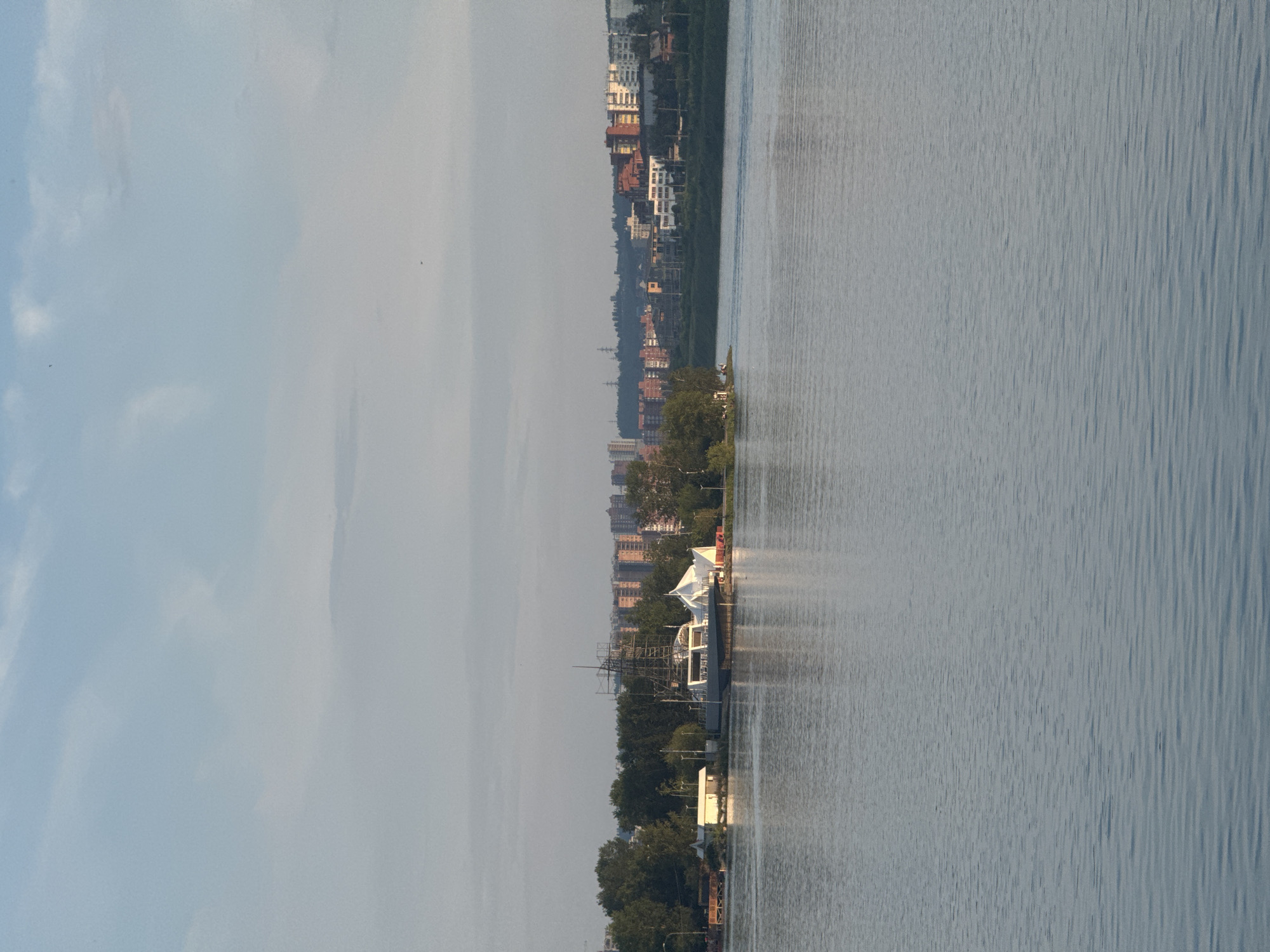 The image depicts a wide body of water, likely a river or lake, with a city skyline in the background. In the foreground, the water is calm with subtle ripples reflecting the overcast sky. On the left bank, there is a small cluster of trees and a distinctive structure that appears to be a ship or boat under construction, surrounded by scaffolding, with a white tent-like structure nearby. Further inland, a dense line of trees separates the water from a collection of multi-story buildings, characteristic of a city. The buildings vary in color and architectural style, suggesting an urban environment. The sky is a pale blue with scattered clouds, indicating a moderately bright day. The overall scene suggests a waterfront area within an urban setting, likely in Irkutsk, Russia, given the context of the request. There are no visible people in the image.