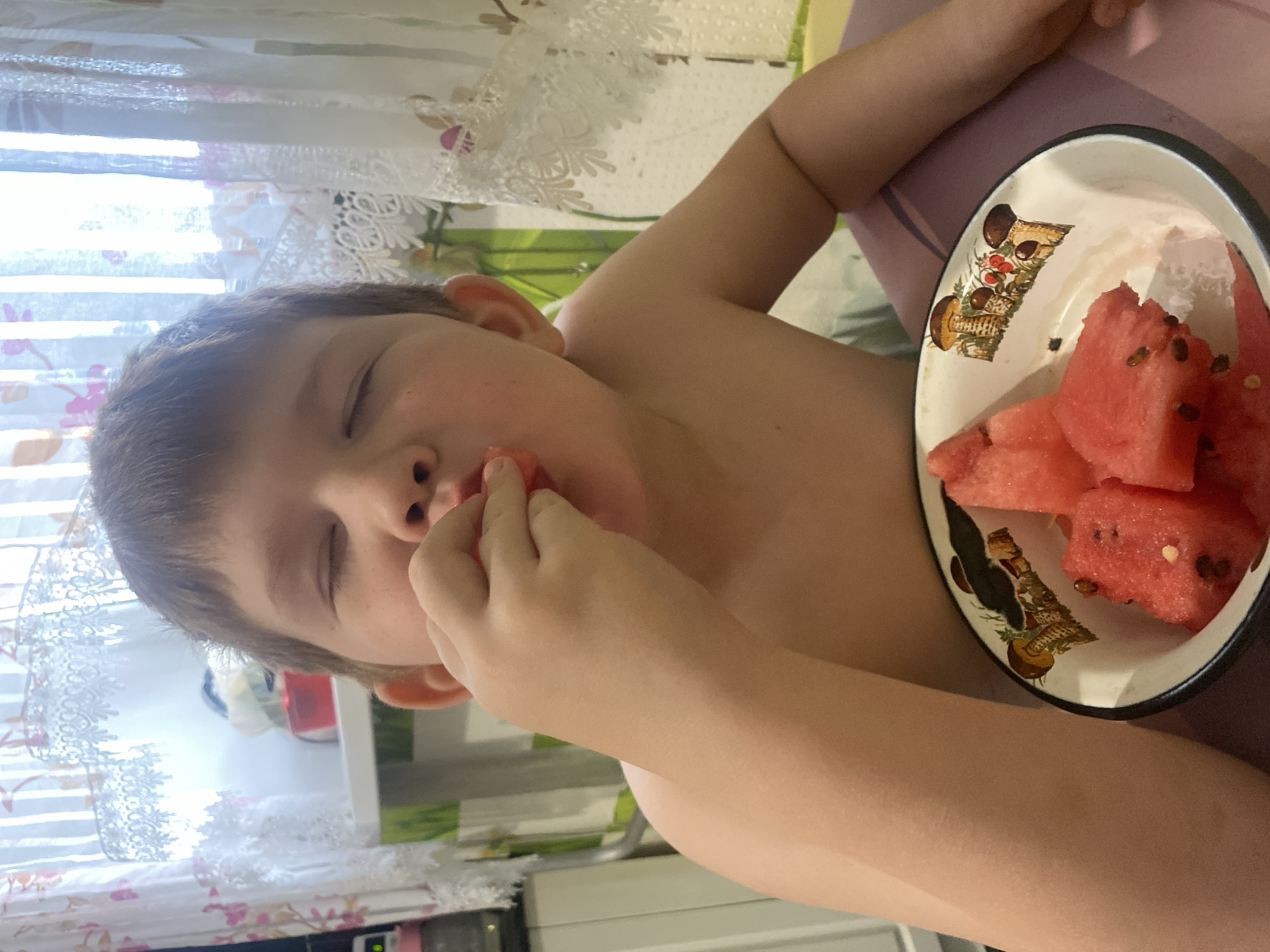 A young boy is eating watermelon from a bowl. The boy is shirtless and sitting at a table. The background shows a window with curtains and some shelves. The scene appears to be indoors, possibly in a home. It is not possible to determine the city of Chaikovsky, Russia, from the image alone.