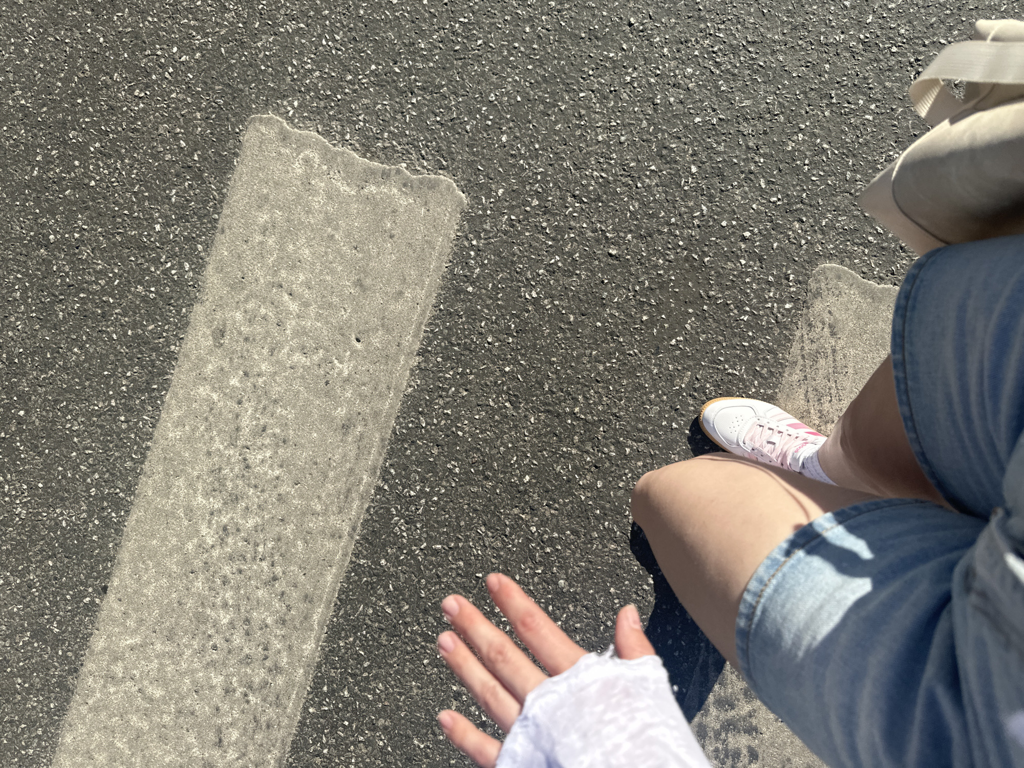 An eye-level shot taken on an asphalt surface, likely a road, features the legs of a person wearing denim shorts and white sneakers with pink laces. A partially visible white object, potentially a hat or bag, is positioned near the upper right corner. The person extends a hand towards the lower left corner, adorned with a white fabric covering. A segment of a worn, light-grey lane marking spans from the upper left towards the center. The photograph does not contain information to determine the location in Perm, Russia.