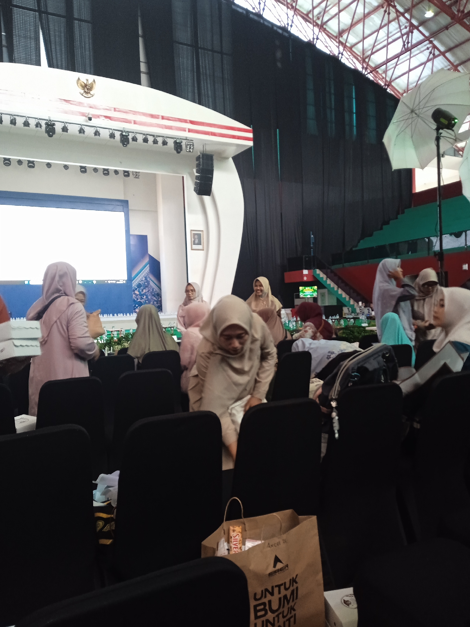The image shows an indoor event, possibly a conference or gathering, taking place in Pangongangan, Indonesia. Several women wearing hijabs are visible; some are standing and talking, while others are seated. In the foreground, black chairs fill the space. A brown paper bag with the text "UNTUK BUMI UNTUK HATI" (FOR EARTH FOR HEART) is partially visible. In the background, a stage with a screen is present, decorated with the Indonesian national emblem (Garuda Pancasila). There's a staircase and seating area in the distance, suggesting the event is in a large hall, perhaps a stadium or auditorium. A photography light with a white umbrella is visible on the right side.