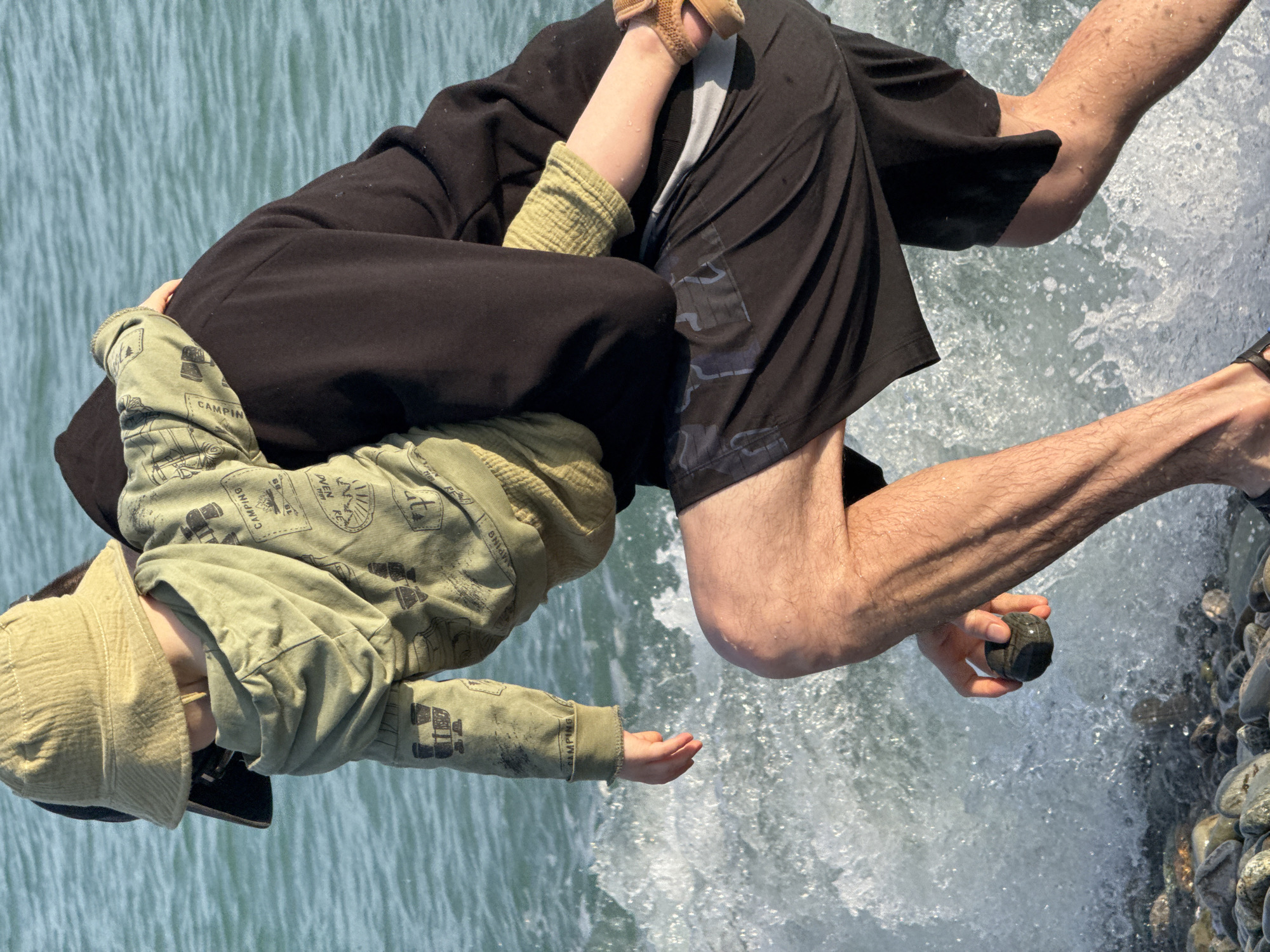 The image shows two people near the water, possibly the Black Sea in Sochi, Russia, though specific landmarks are not visible. A fair-skinned adult, wearing black shorts and sandals, is holding a smooth stone. A child, wearing a green patterned jacket and a bucket hat, is either being held or leaning against the adult. The background consists of water, pebbles, and potentially a shoreline. The water is splashing, suggesting some movement or waves.