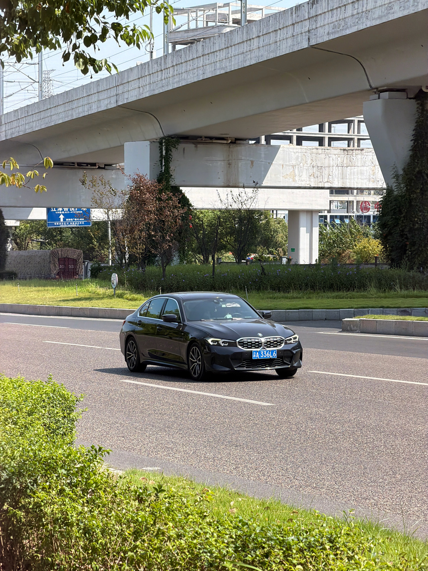 A black BMW sedan, license plate A 336L6, is driving on a paved road with a white lane marking. The car is positioned in the right lane, facing forward and slightly to the left. In the background, there is an elevated concrete structure, likely an overpass or bridge, with visible support pillars. Trees and greenery flank the road and the area beneath the overpass. A blue traffic sign with Chinese characters and an arrow pointing upwards is visible on the left side of the road. Sunlight is casting shadows. The scene appears to be in an urban or suburban environment in Jiangjin, China.