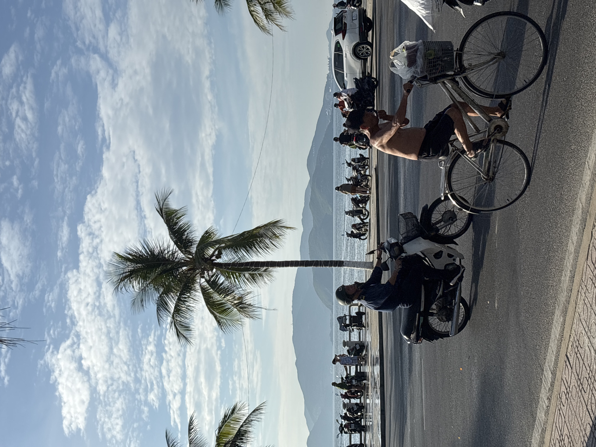 In Nha Trang, Vietnam, under a sky with scattered clouds, a man rides a bicycle along a paved road. He is shirtless, wearing dark shorts and sandals, and is smoking. The bicycle has a basket on the front containing a plastic bag. Behind him, another man wearing a dark uniform and a helmet rides a scooter. The road is bordered by a sidewalk with a patterned tile surface. In the background, several motorcycles are parked along a promenade adjacent to the ocean. The ocean stretches to a mountainous coastline under the bright sky. A white SUV is also visible parked in the distance. Palm trees line the area, with their fronds reaching into the sky.